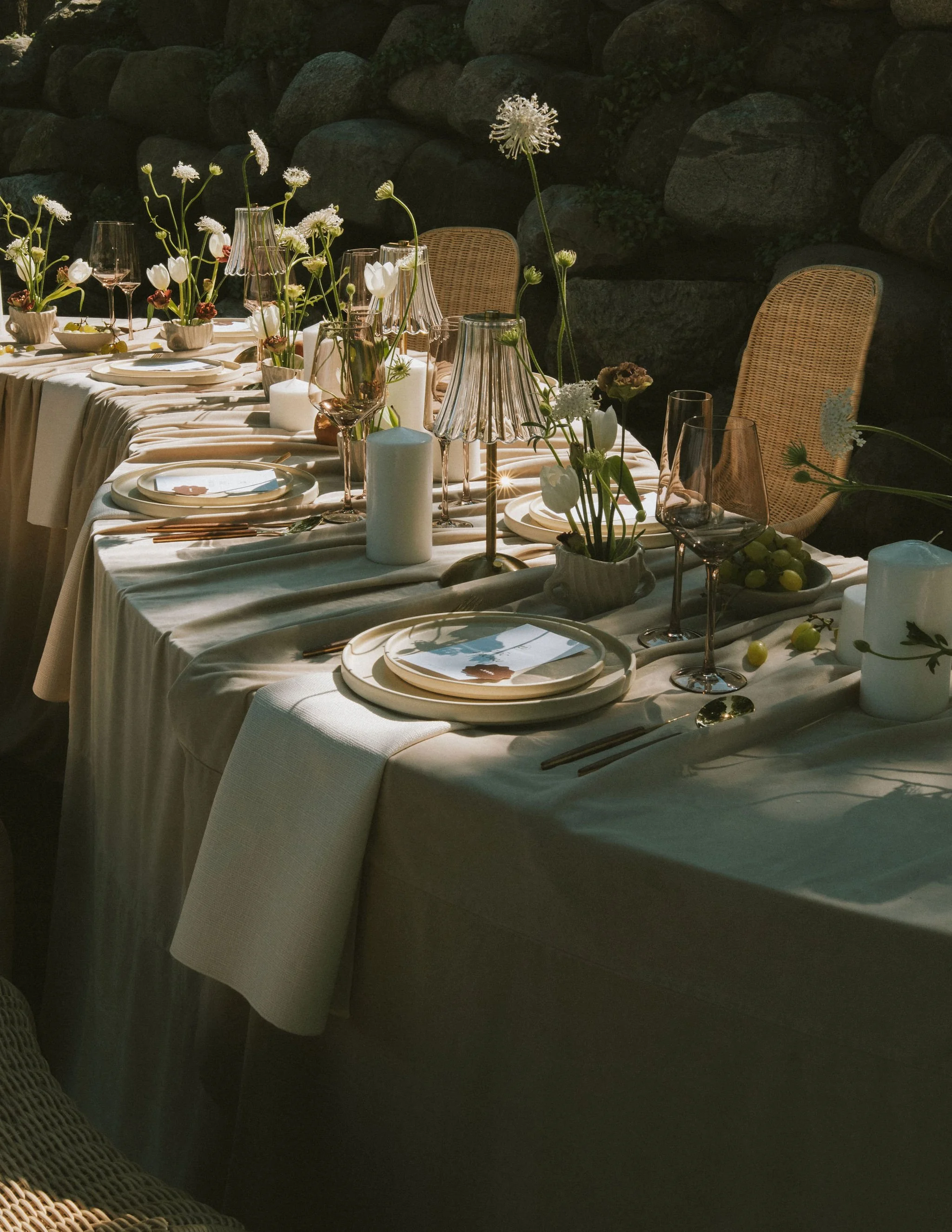 A beautifully set outdoor dinner table with white tablecloth, floral centerpieces, candles, wine glasses, and place settings, with chairs and a rock wall in the background.