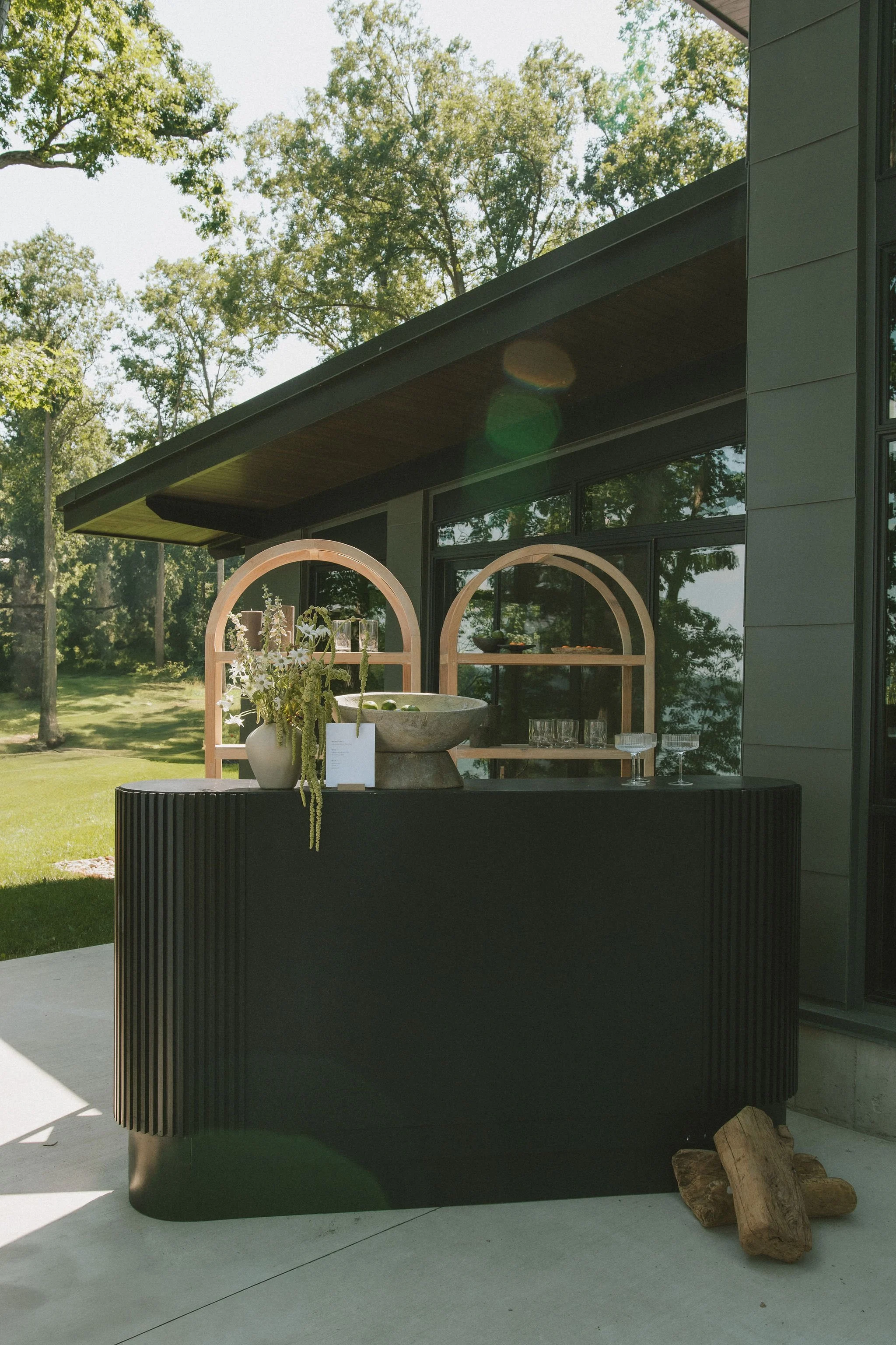 Outdoor bar setup with a black curved bar counter, a large stone bowl, a vase with flowers, and two wooden arch frames, set against a modern house with large windows and surrounded by trees.