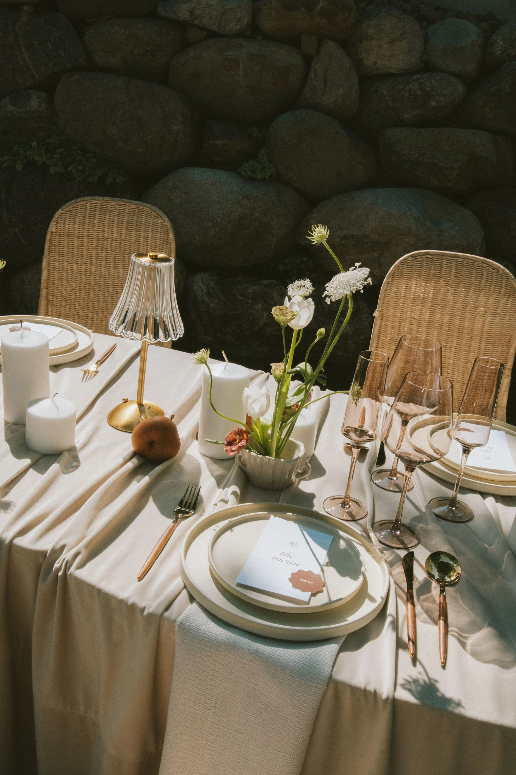 A dining table set with white plates, glasses, and cutlery, decorated with a flower arrangement, candles, and a small lamp, against a stone wall backdrop.