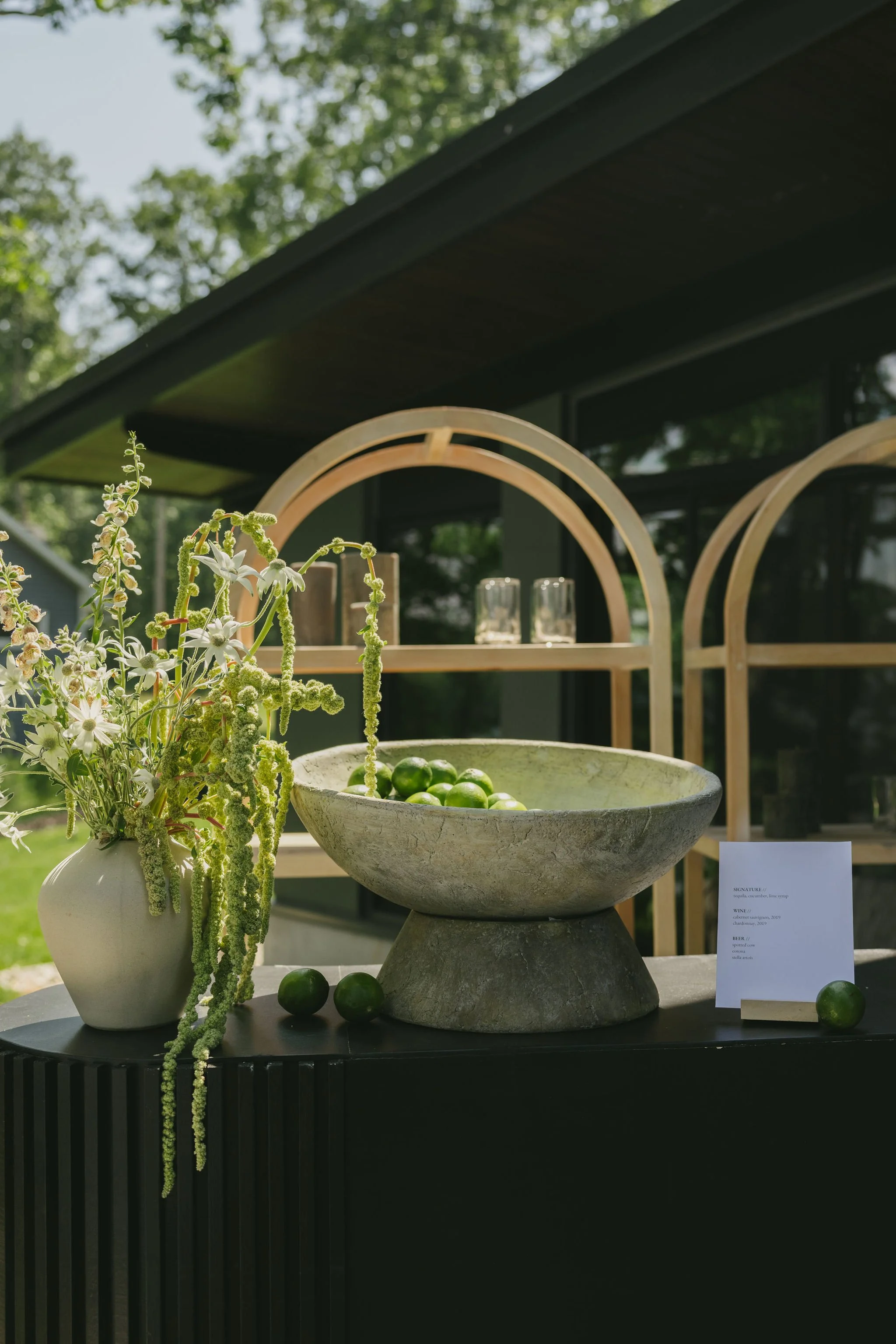 A decorative outdoor display with a beige vase of white and green flowers, a large stone bowl filled with green limes, and a small white sign on a black table. In the background, there are three wooden arches and glassware, with a house and trees visible beyond.