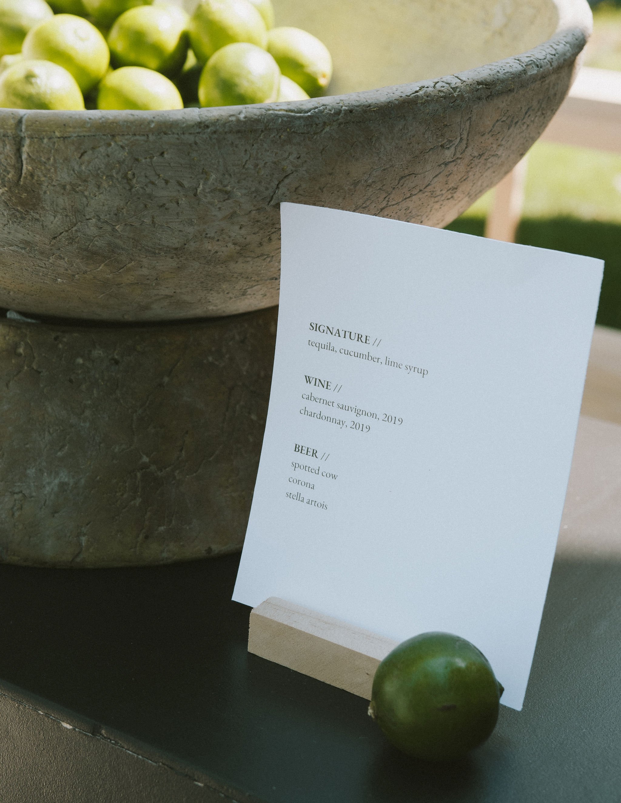 A bowl of green limes, a white menu card listing drink options, and a single lime placed in front of the menu card on a dark surface.