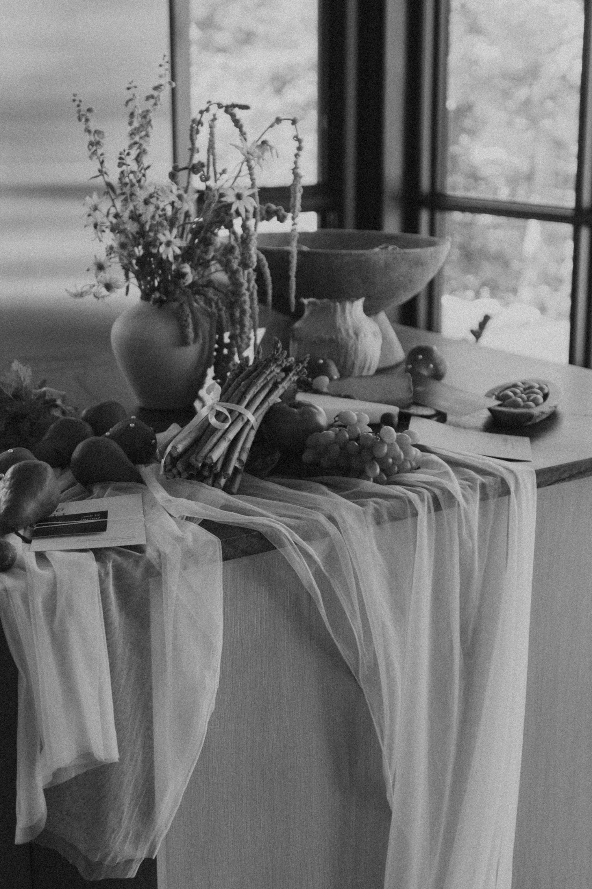 Black and white photo of a table decorated with fruits, flowers, and a cloth, set near a large window with outdoor scenery.