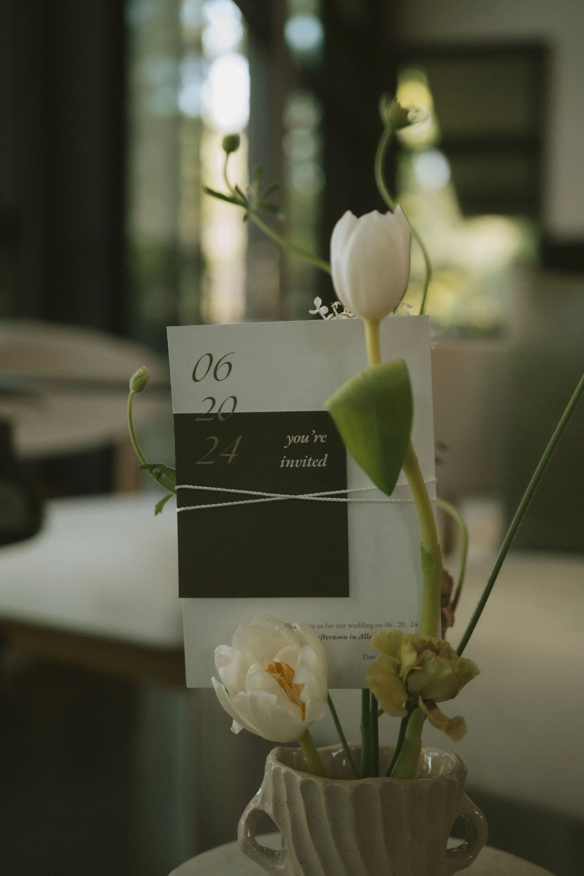 A floral arrangement with white and green flowers holding a black and white wedding invitation card with the date 06.20.24 and the text 'you're invited'. The arrangement is in a beige or light brown ceramic vase on a table, with a softly blurred background of a window and indoor space.