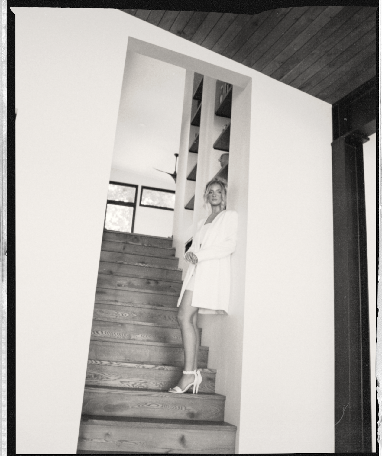 A woman standing on wooden stairs inside a modern house, wearing high heels and a blazer.