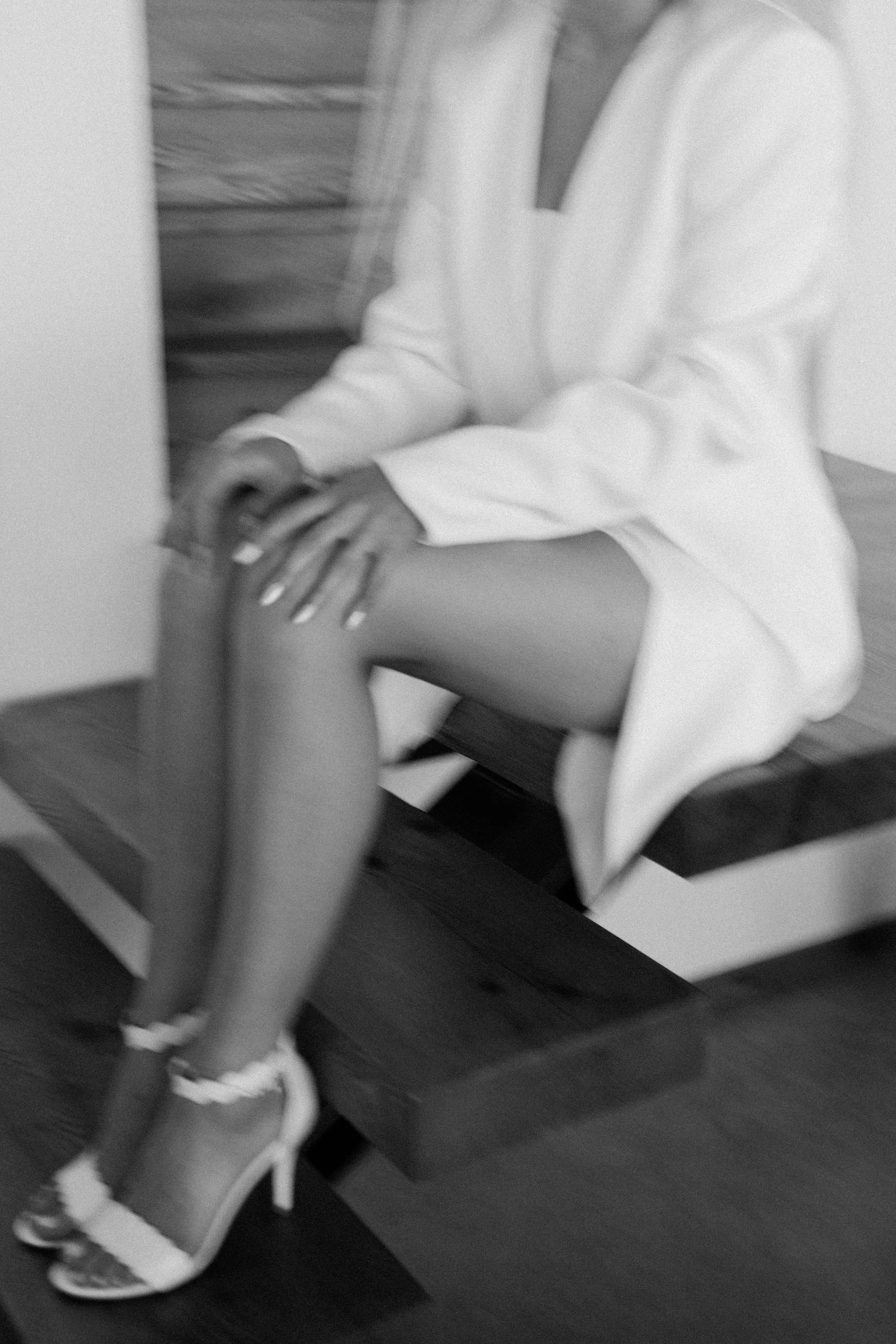 Black and white photo of a woman sitting on a bench, wearing high-heeled shoes, a dress, and a light-colored blazer, her hands resting on her knee.