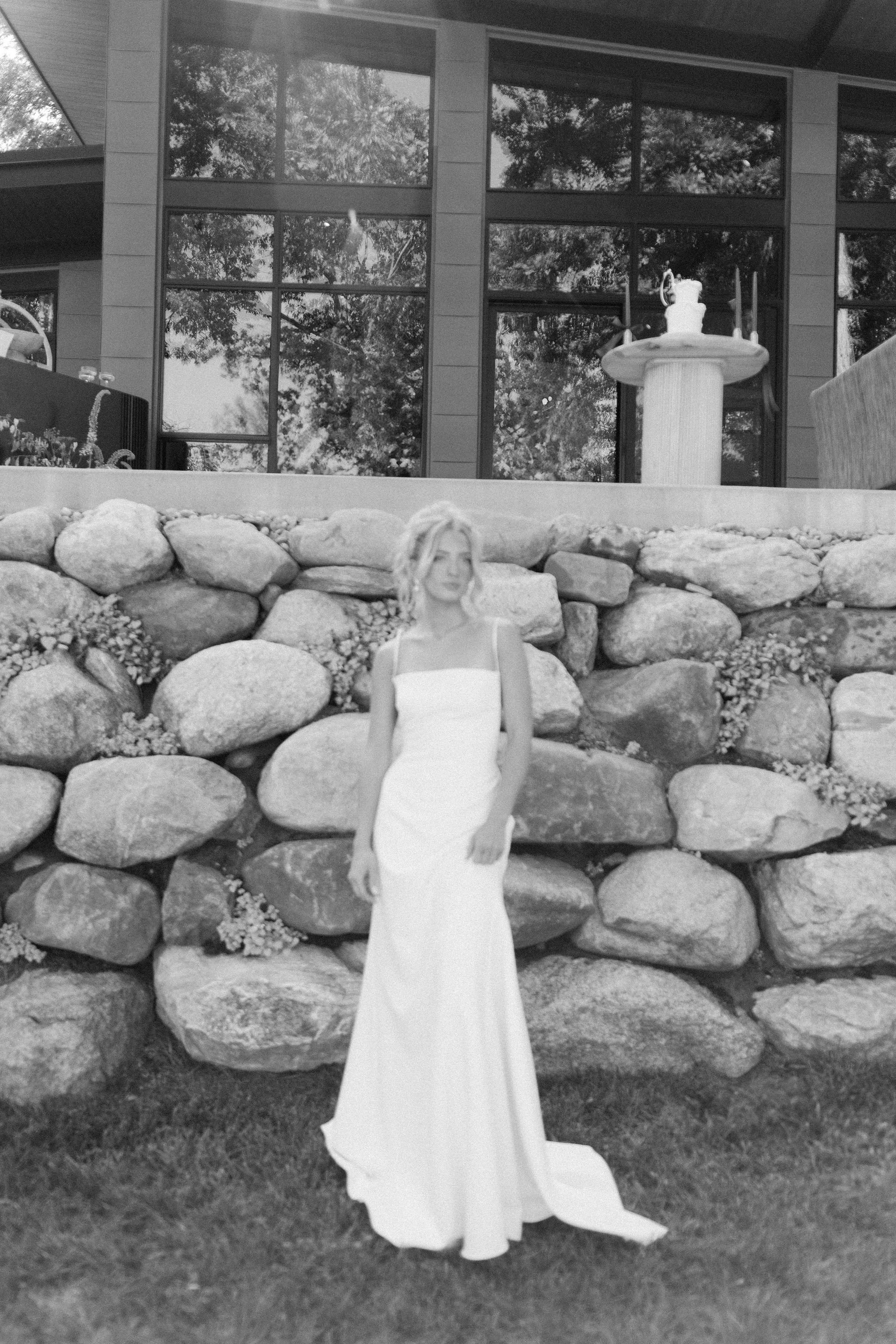 A woman in a white dress standing outdoors in front of a stone wall, with a modern building and large windows behind her.