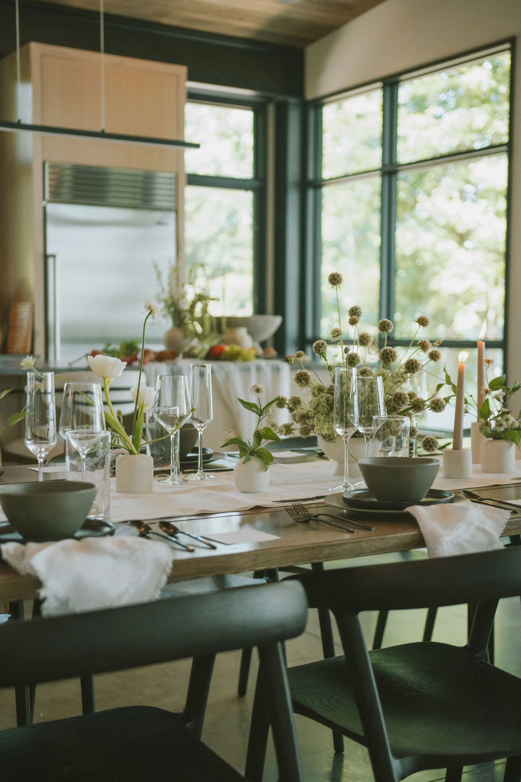 A dining table set for a meal with white tablecloth, green and white flower arrangements, candles, and black plates and cutlery, with large windows and a view of greenery outside.