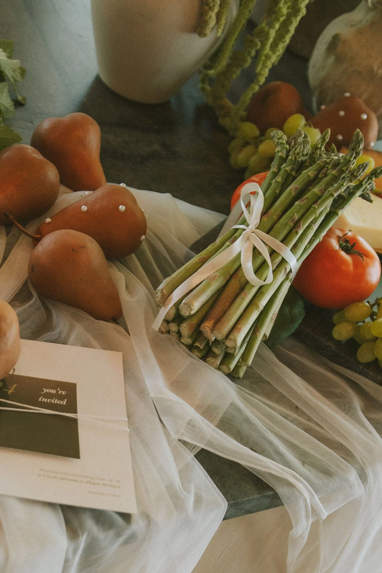 A bundle of fresh asparagus tied with a white ribbon, surrounded by tomatoes and apples on a table decorated with white sheer fabric.