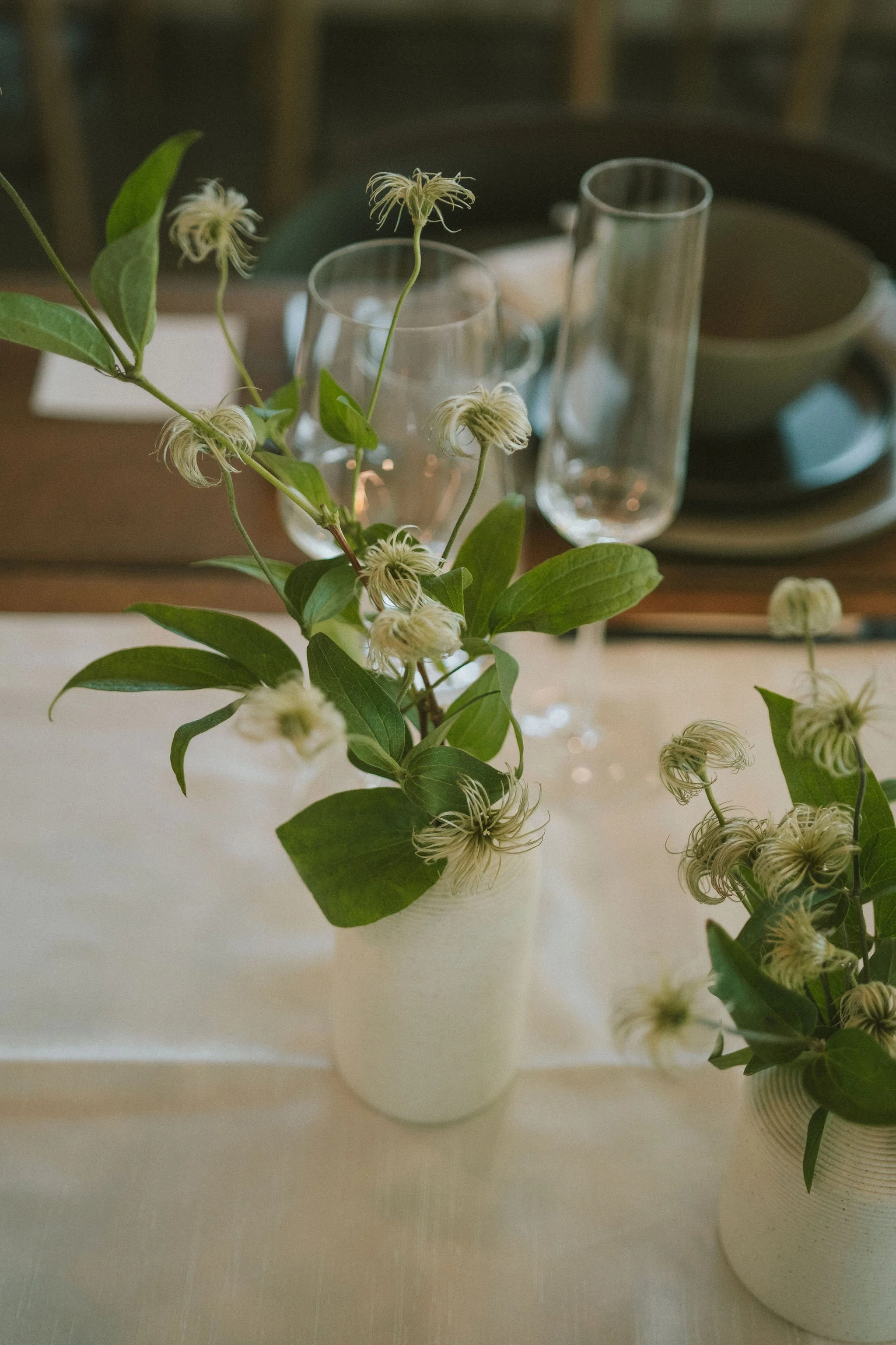 A table centerpiece with a white vase of green leaves and white flowers, surrounded by empty glasses and dinnerware.