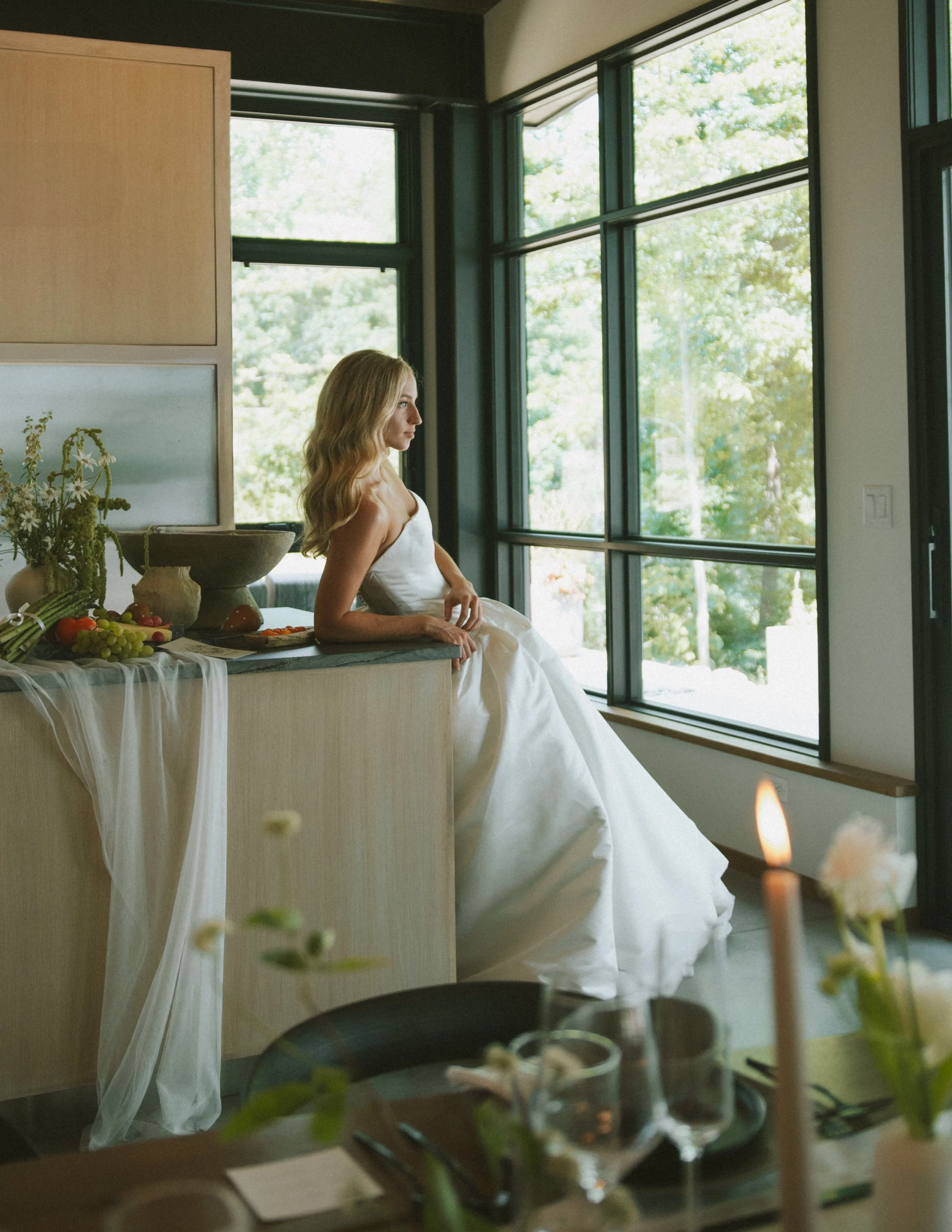 A woman in a white strapless dress sitting on a kitchen counter near large windows, overlooking a green outdoor scene.