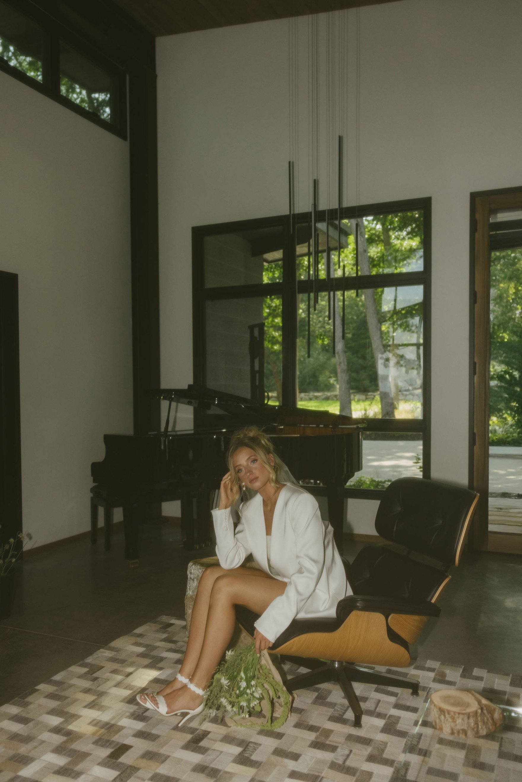 A woman sitting in a modern living room with large windows, a grand piano in the background, and contemporary lighting fixtures hanging from the ceiling. She is dressed in white, sitting on a wooden chair with a patterned rug beneath her, and is holding a small bouquet of flowers.