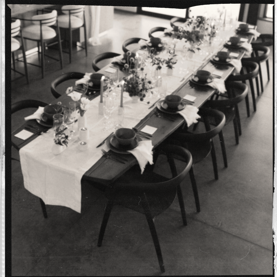A long dining table set for a meal with black bowls, glasses, and cutlery, decorated with floral arrangements and candles, in a black and white photo.