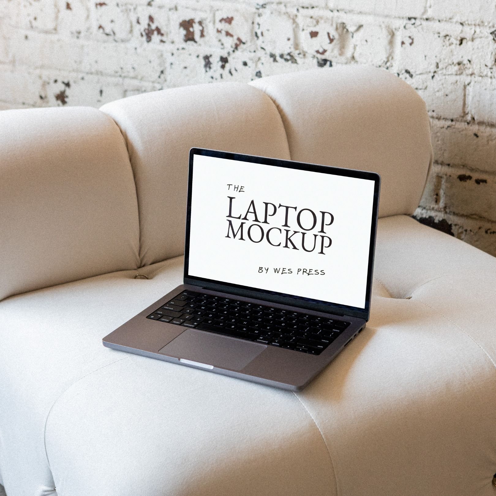 A laptop resting on a white cushioned sofa, displaying a screen with the text 'The Laptop Mockup by Wes Press' against a brick wall background.