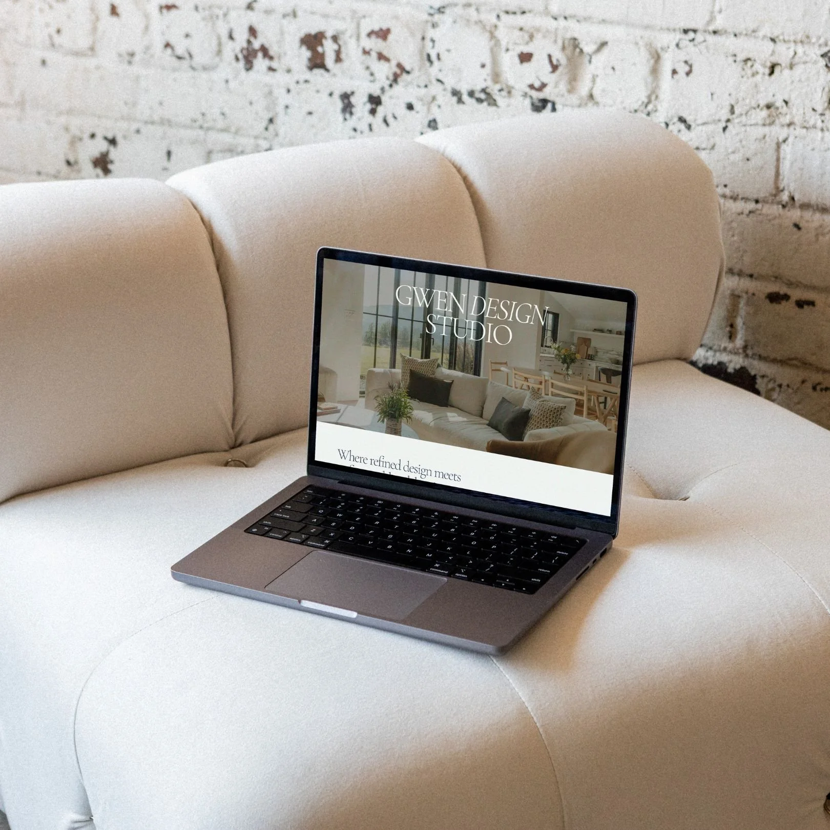 A laptop placed on an off-white cushioned sofa with a textured brick wall in the background. The laptop screen displays a website for Gwen Design Studio with an interior image and text.