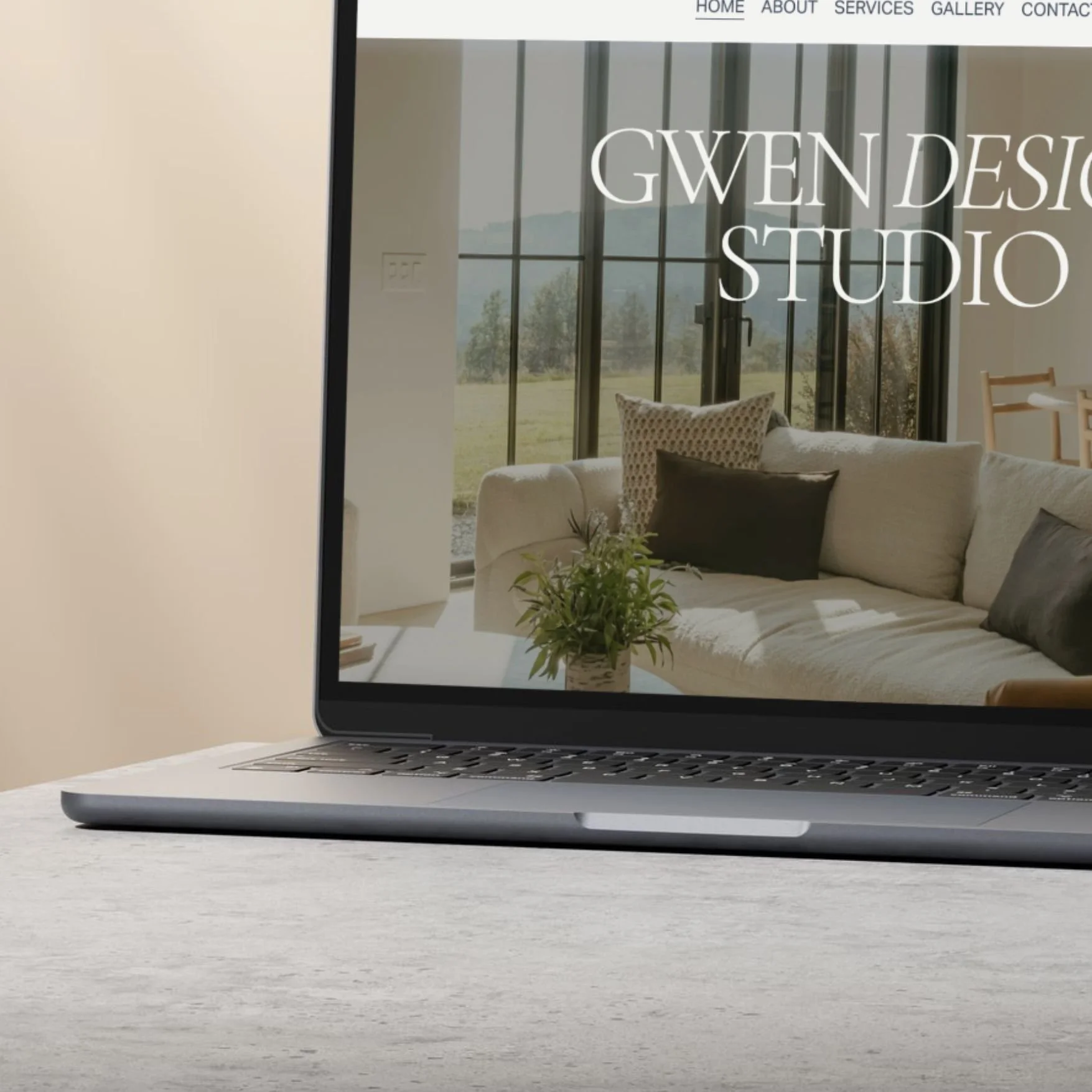 A laptop on a light-colored table displaying a website for Gwen Design Studio, showing an interior living room with a beige sofa, black and white pillows, a potted plant, and large windows overlooking an outdoor landscape.