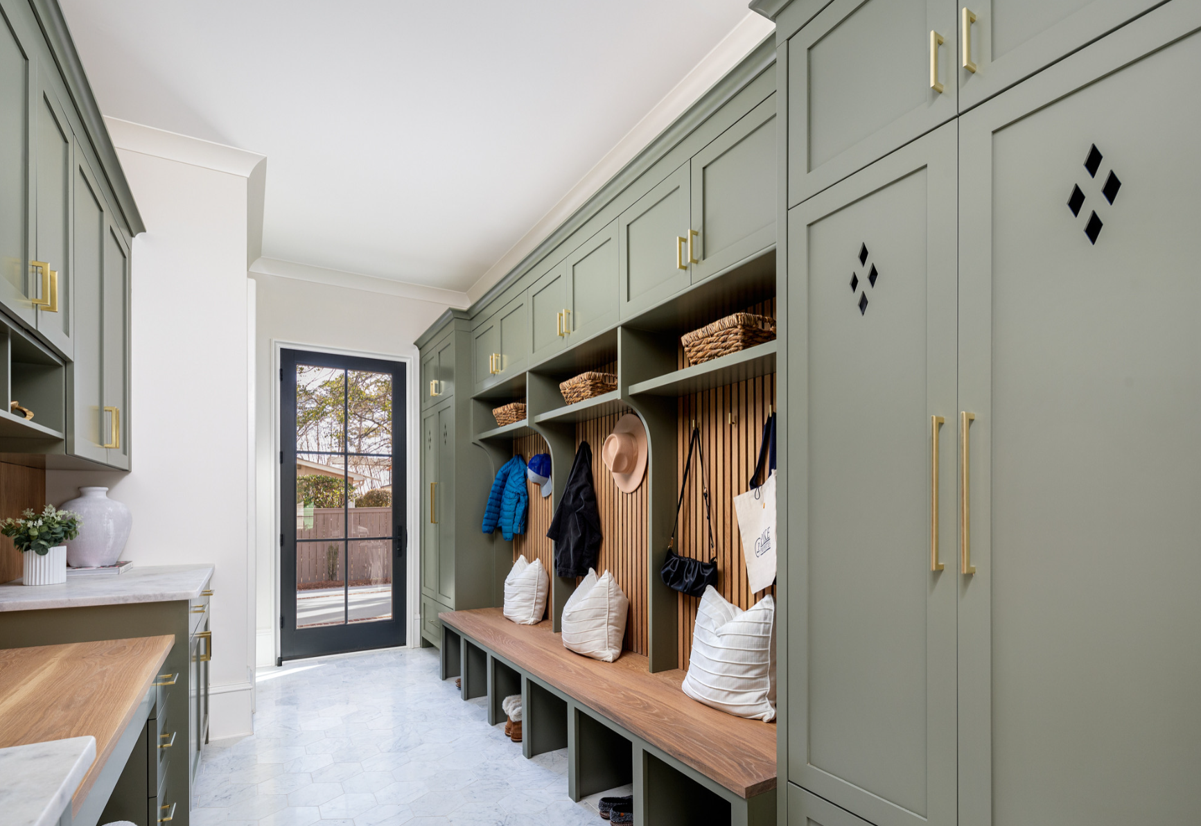 Mudroom with green built-in cabinets, wooden bench with pillows, hooks for coats and hats, baskets on top shelves, and a door leading outside with a view of a backyard.