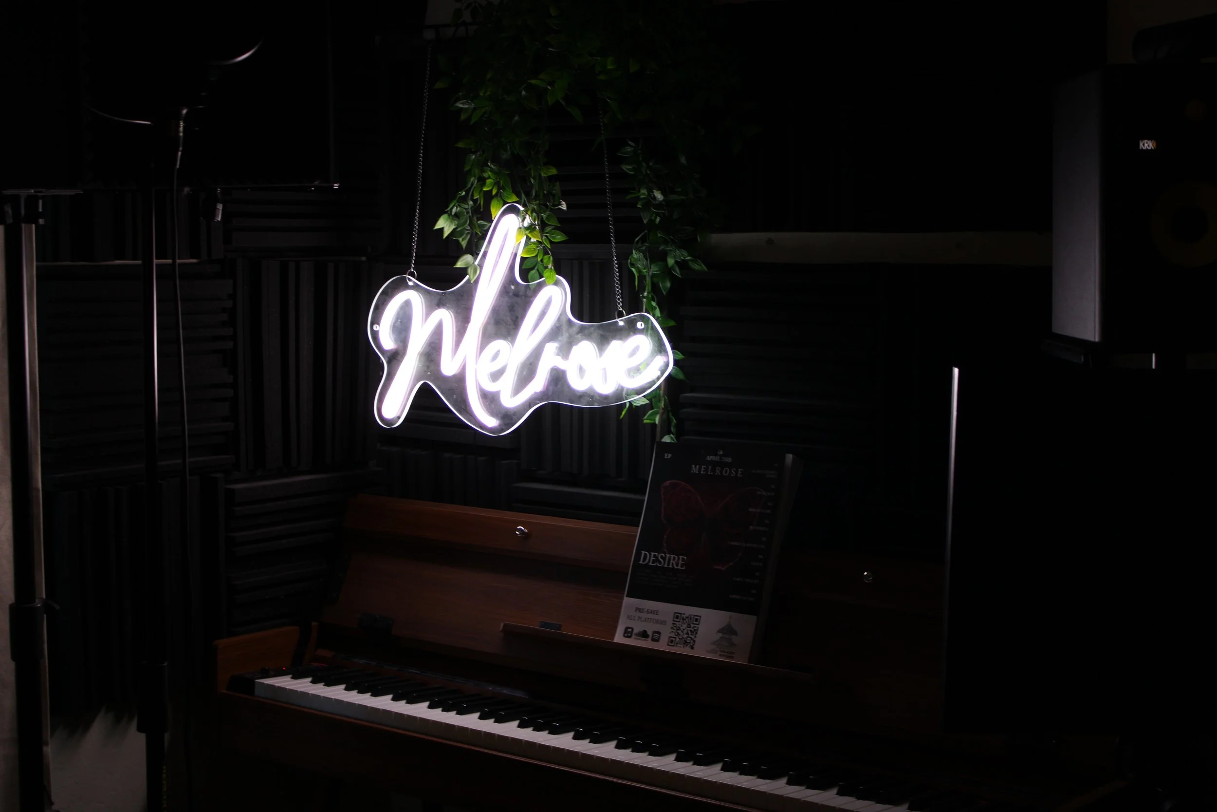 A neon sign spelling 'hello' hangs above a wooden piano with a sheet of music on its music stand, surrounded by dark interior and greenery.