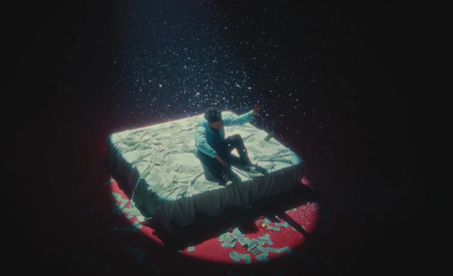 A person sitting on a bed surrounded by scattered paper money, illuminated by a spotlight against a dark background with floating particles resembling stars.