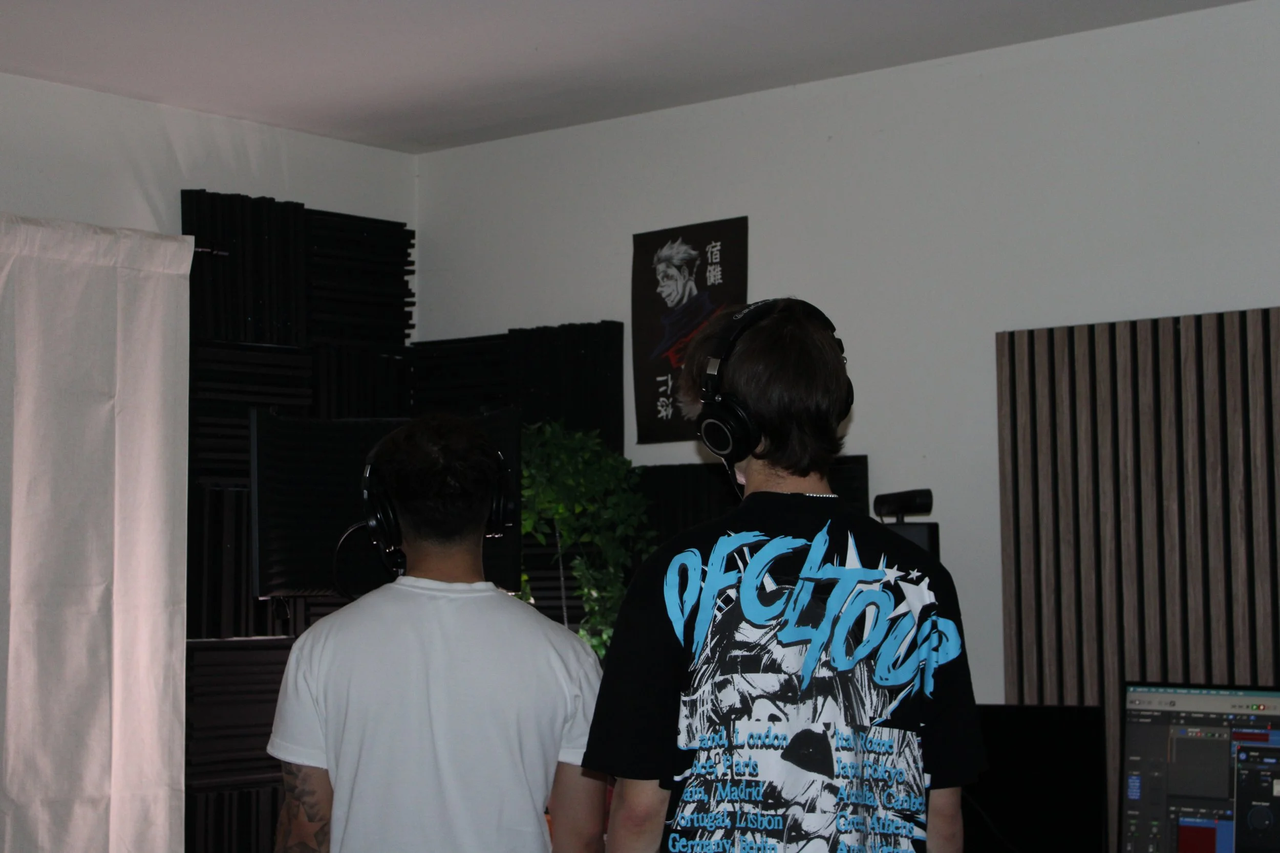 Two individuals with headphones working at a music studio, facing away from the camera, with soundproofing panels and a poster on the wall.