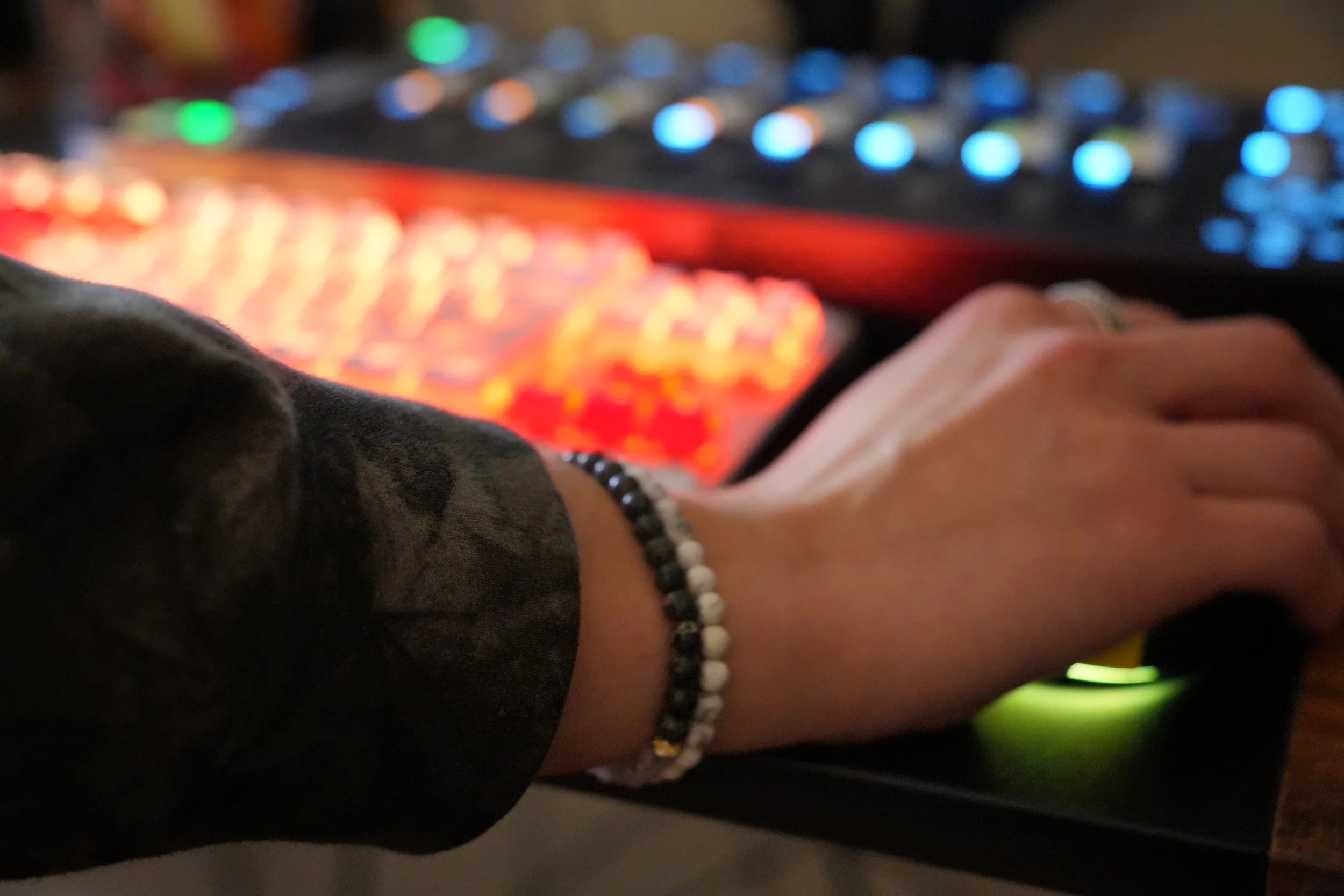 Close-up of a person's hand with a beaded bracelet, in front of a blurred background of colorful lights, possibly a DJ or music setup.