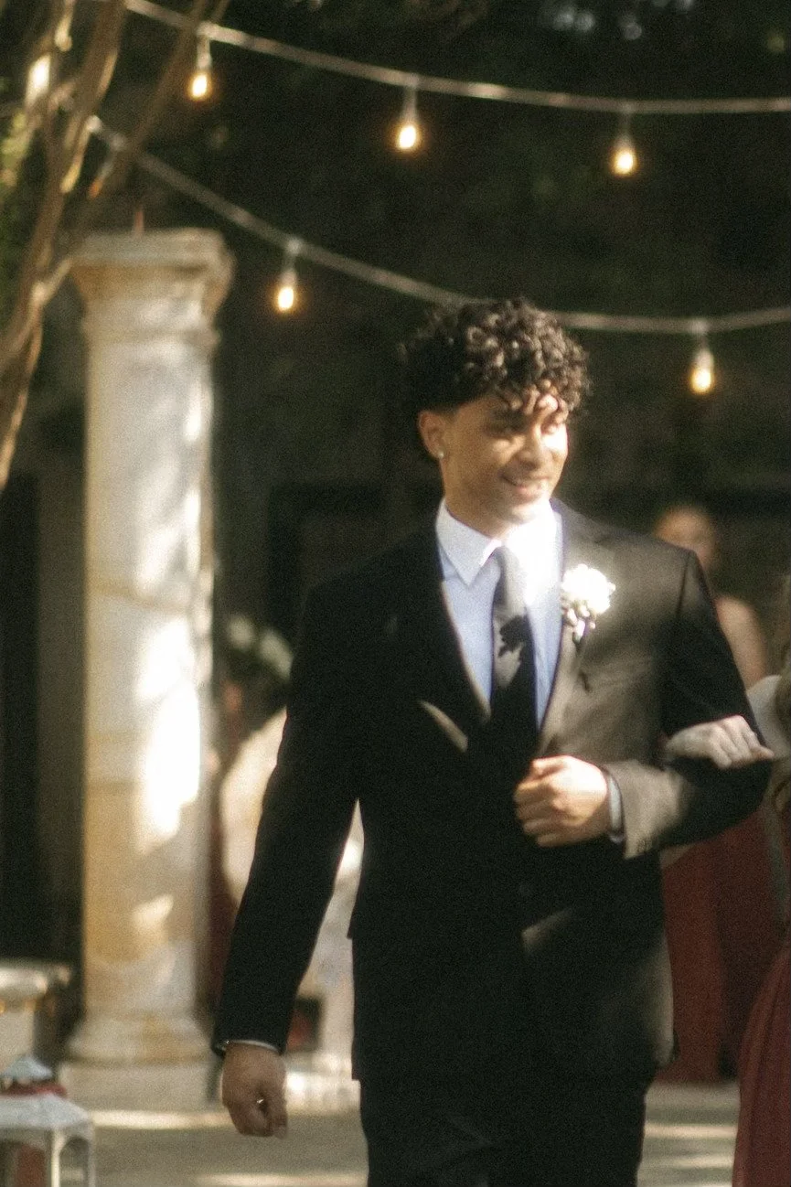 A smiling man in a black suit, white shirt, and tie, standing outdoors under string lights, in a festive setting.