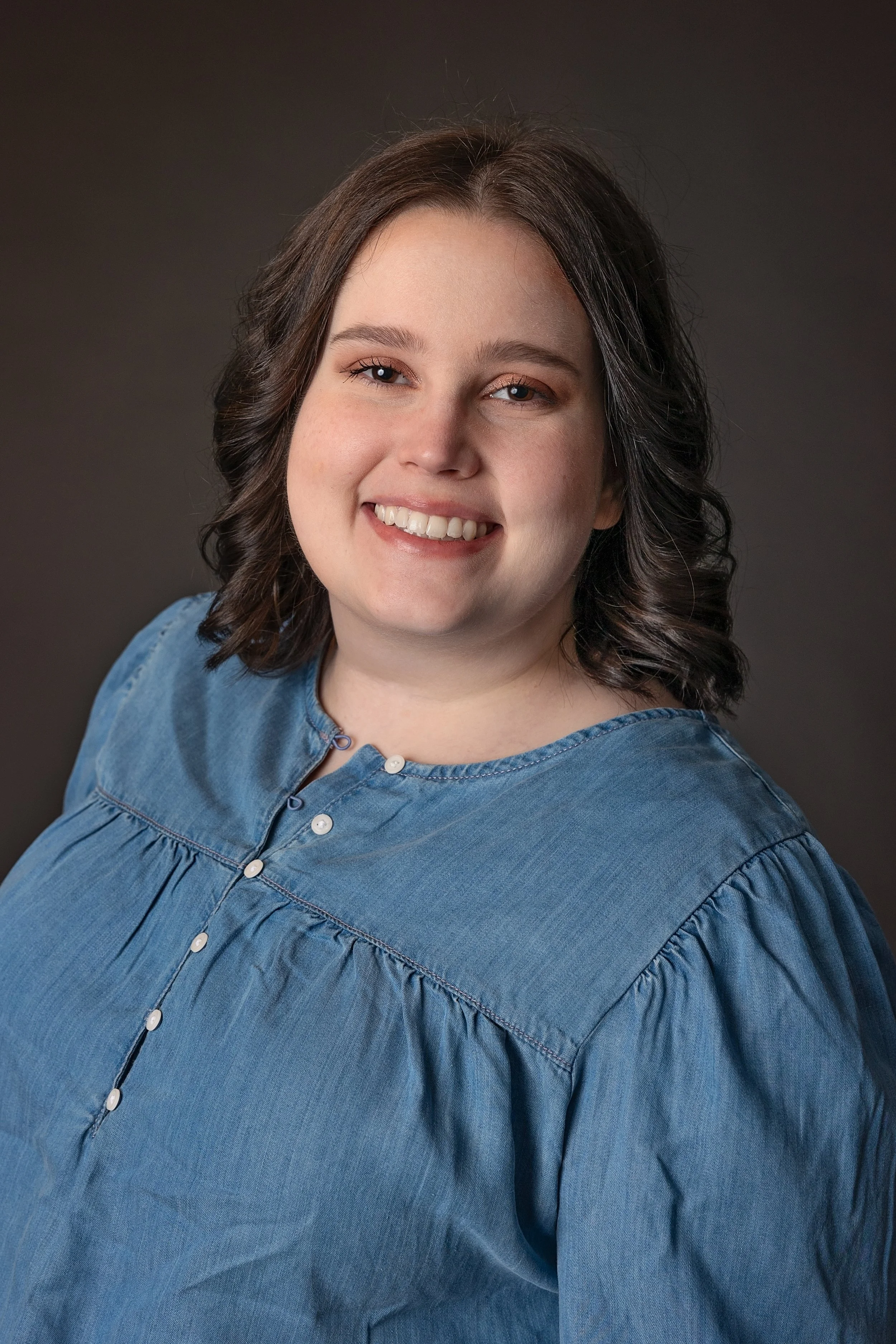 Close-up of a young woman with shoulder-length brown hair, smiling, wearing a blue denim button-up shirt, against a dark gradient background.