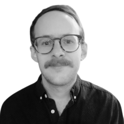 A young man with glasses and a mustache, wearing a dark button-up shirt, smiling at the camera. Black and white photo.