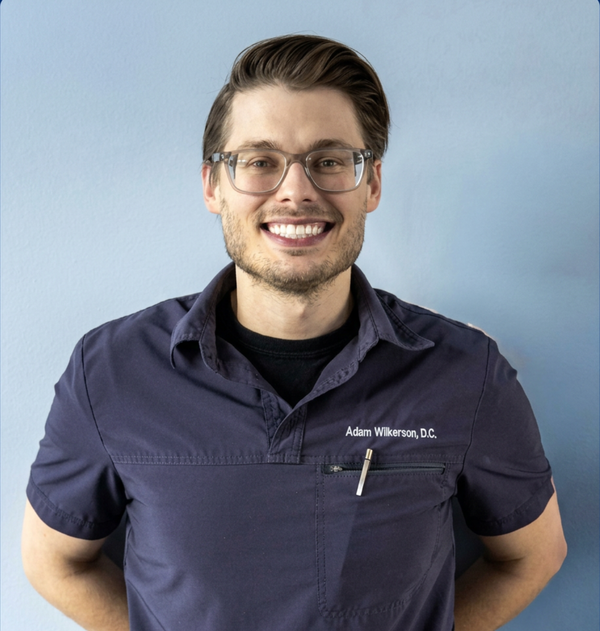 Professional man with glasses and a dark shirt, smiling at the camera, standing against a light blue wall.
