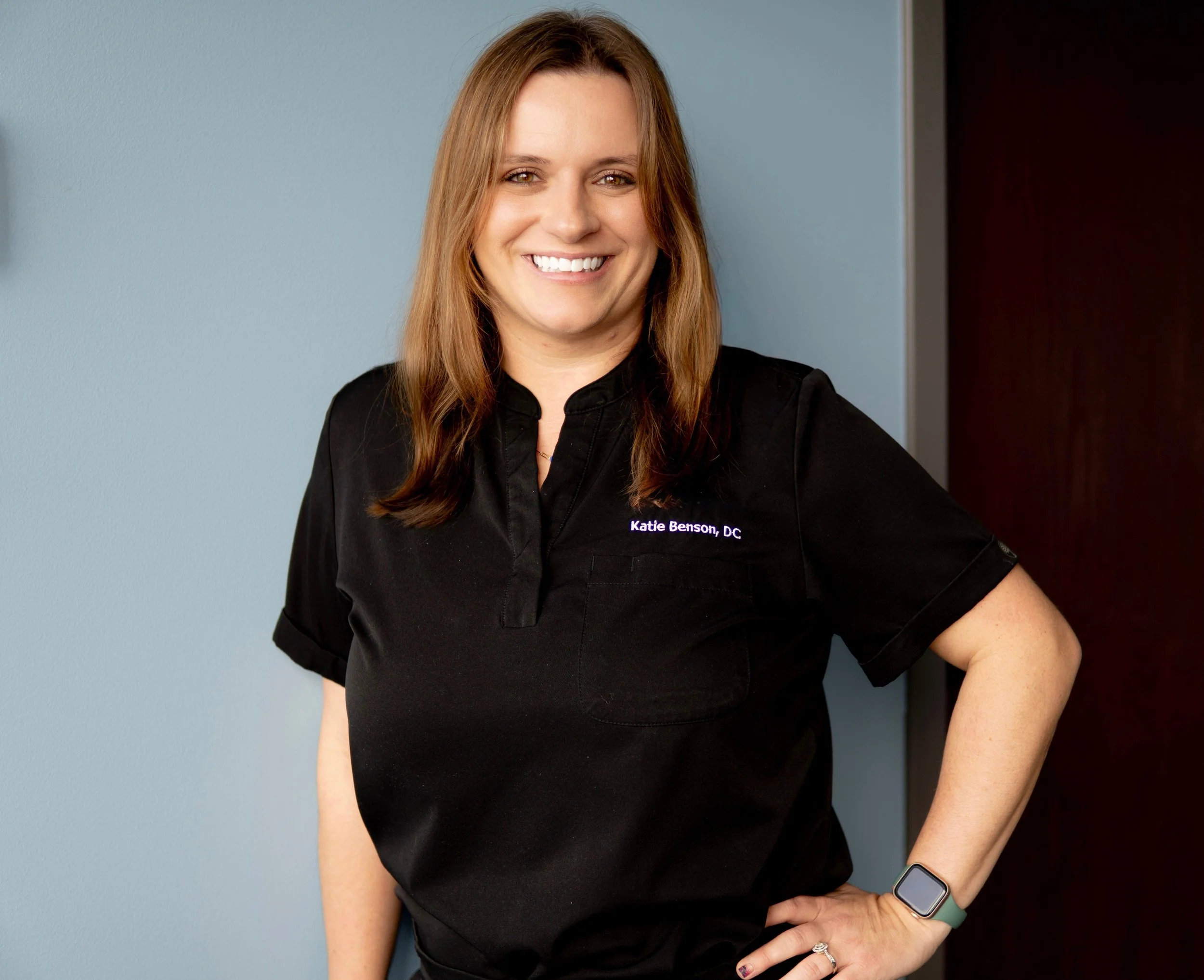 A woman with shoulder-length light brown hair, smiling, wearing a black top with the name tag 'Katie Benson, DC'. She has a smartwatch on her left wrist and multiple rings on her fingers, standing against a blue wall with a dark wooden door partially visible on her right.