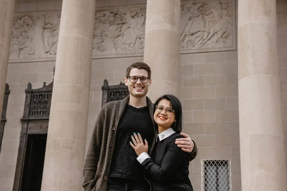 A smiling man and woman stand together in a building with tall stone columns and decorative carvings. The man has short brown hair, glasses, and is wearing a dark sweater and a brown cardigan. The woman has black hair, glasses, and is dressed in a black jacket with a white collar. They are embracing with the woman showing a ring on her finger.