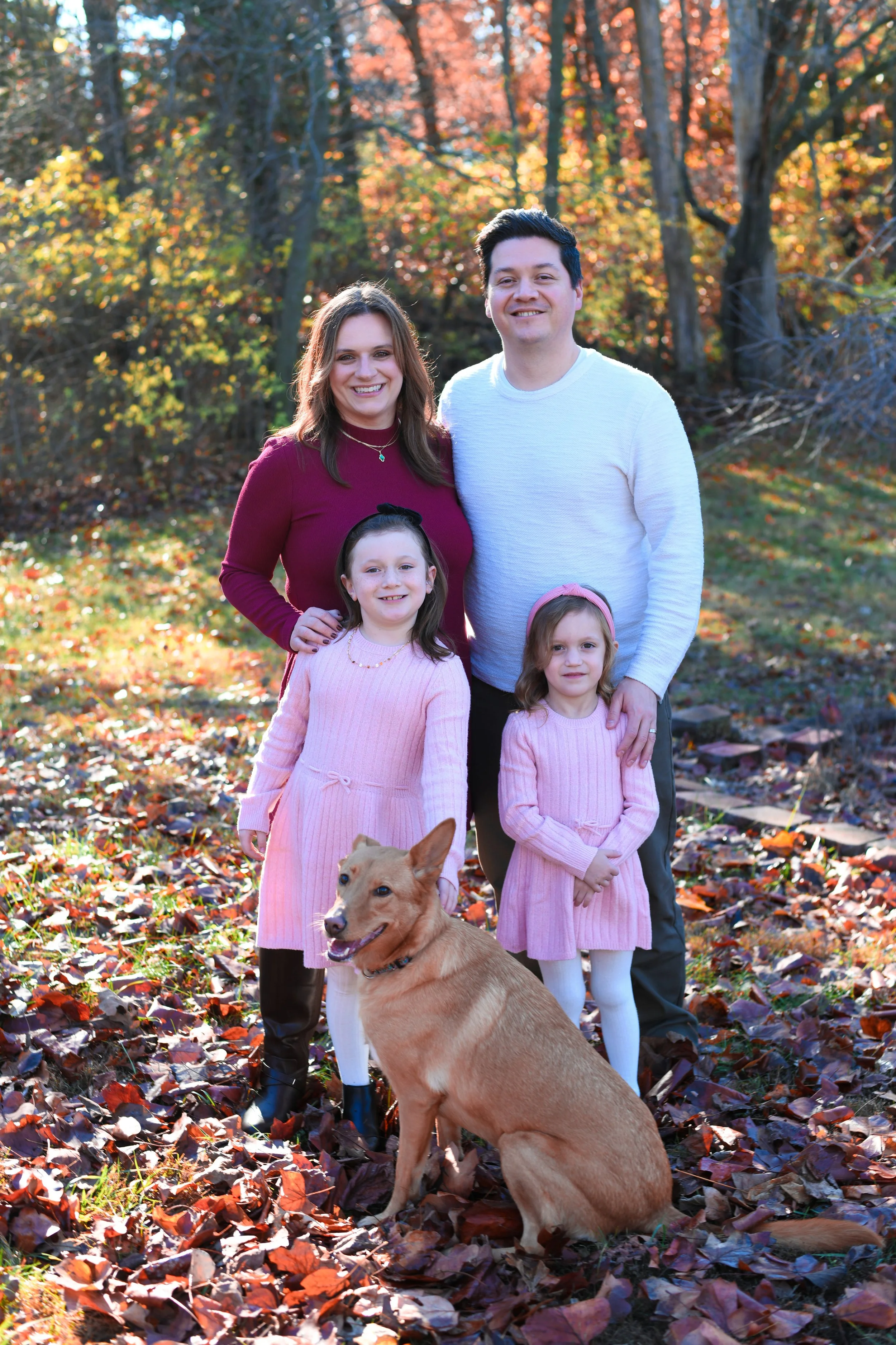 A family of four with a dog posing outdoors in an autumn setting with colorful fall leaves on the ground and trees in the background.