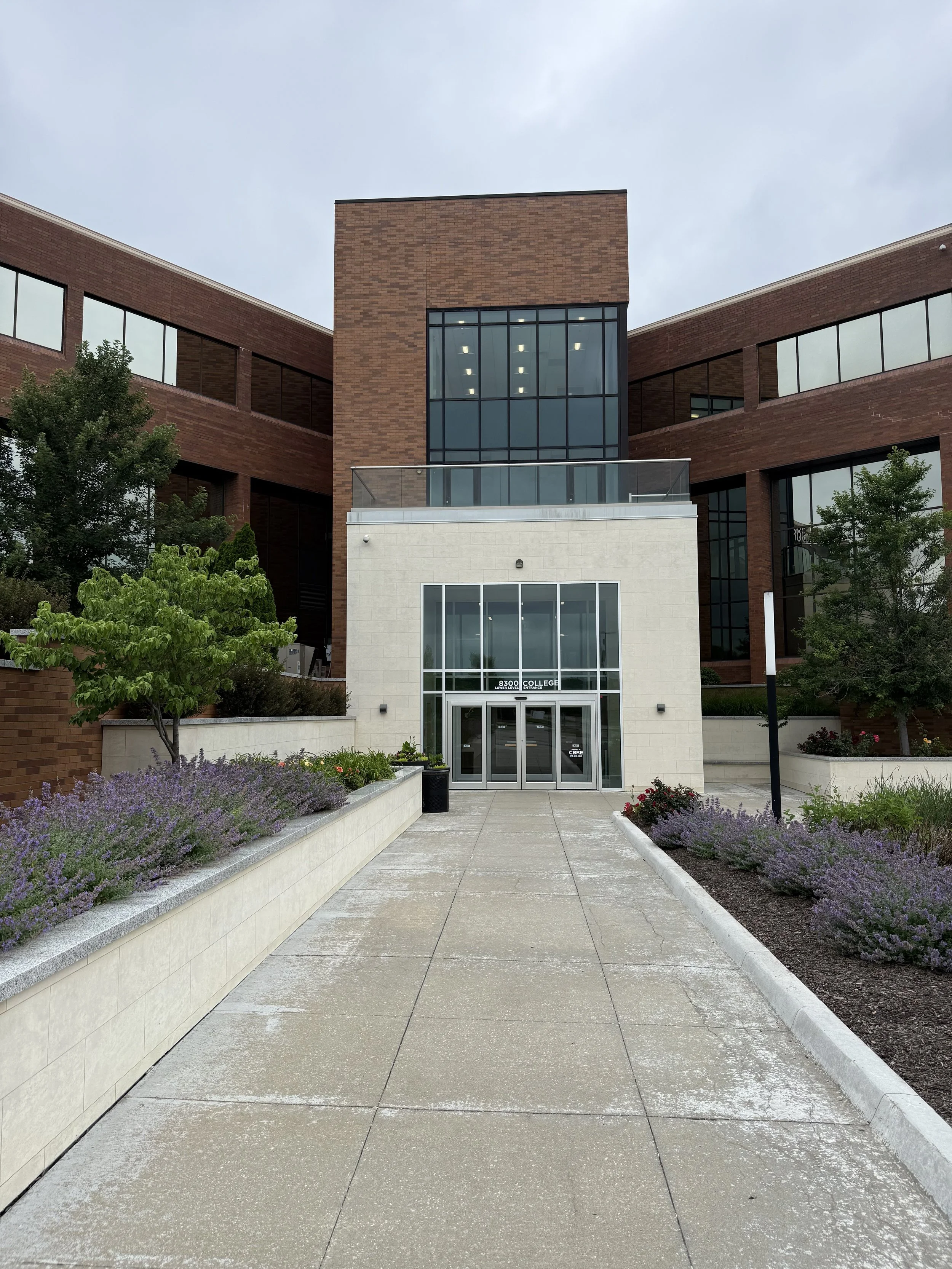 The entrance to a college building with the address 8300 written above the doors, and landscaping along the walkway leading to the front door.