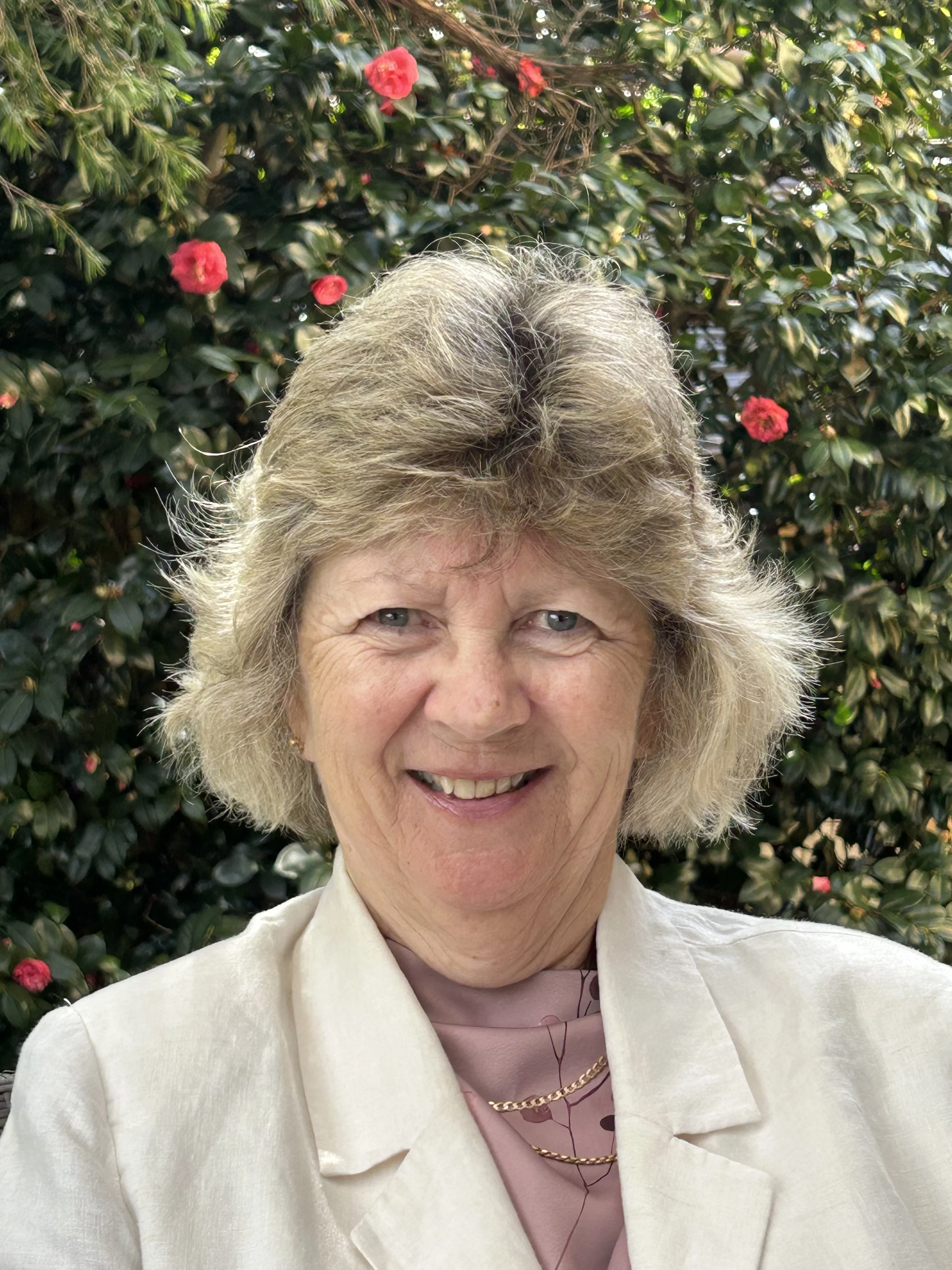 An elderly woman with gray curly hair smiling, wearing a light cream blazer and a pink top with a floral pattern, standing in front of green bushes with pink flowers.