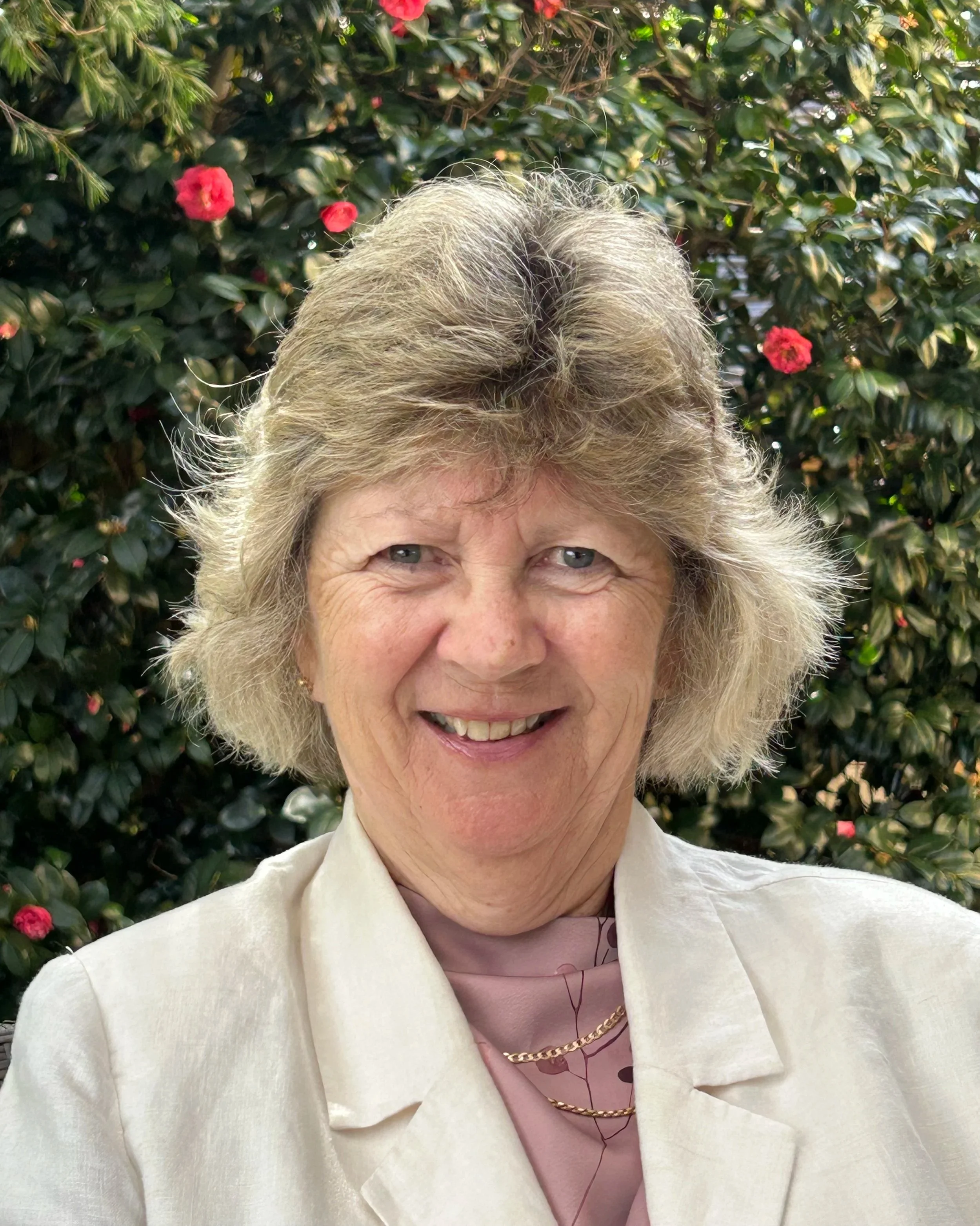 Close-up of an older woman with gray hair and light skin, smiling, outdoors with green foliage and pink flowers in the background.