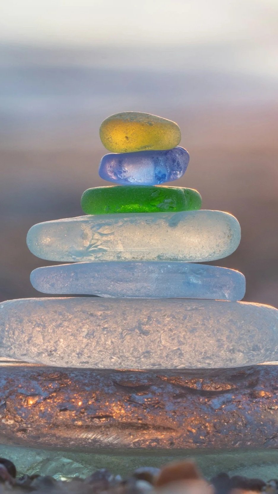Stacked colorful glass stones, with blue, green, yellow, and clear stones, set against a blurry background with warm sunlight.