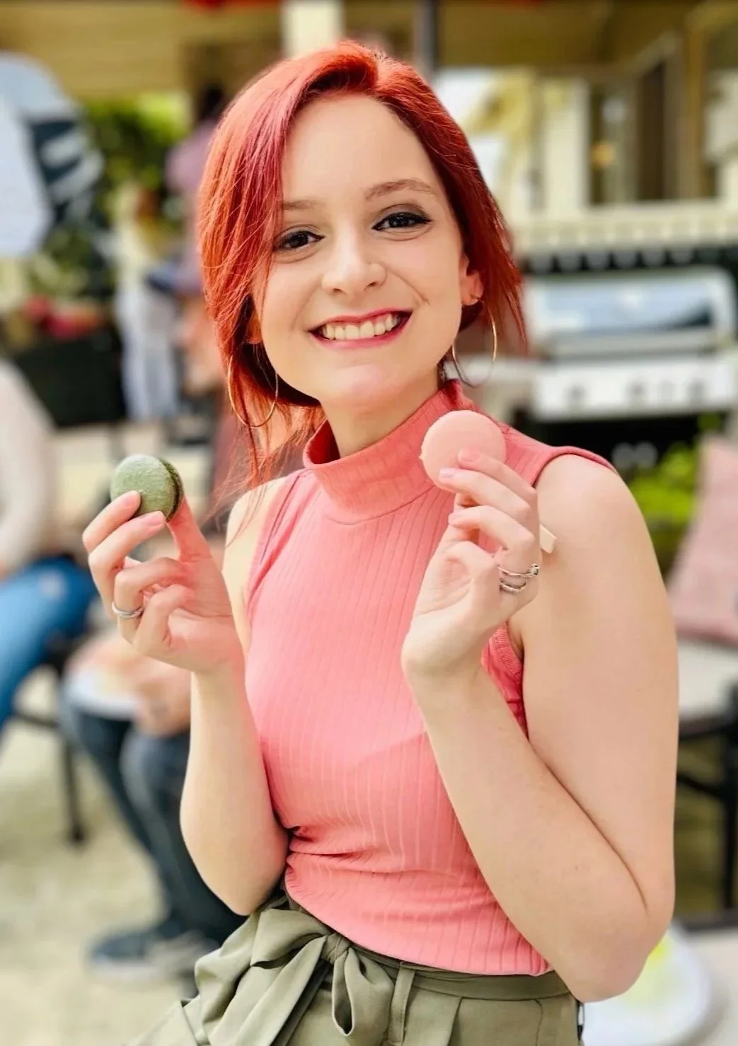 A young woman with red hair smiling and holding a macaron in each hand, wearing a sleeveless pink top and beige pants, outdoors at a social gathering.