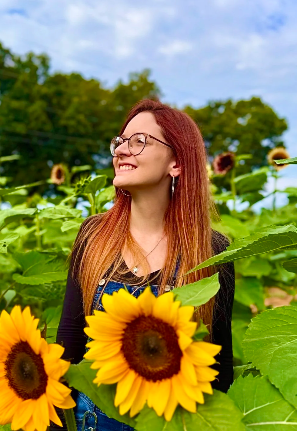 A woman with red hair and glasses smiling while standing among tall sunflowers in a sunflower field during daytime.