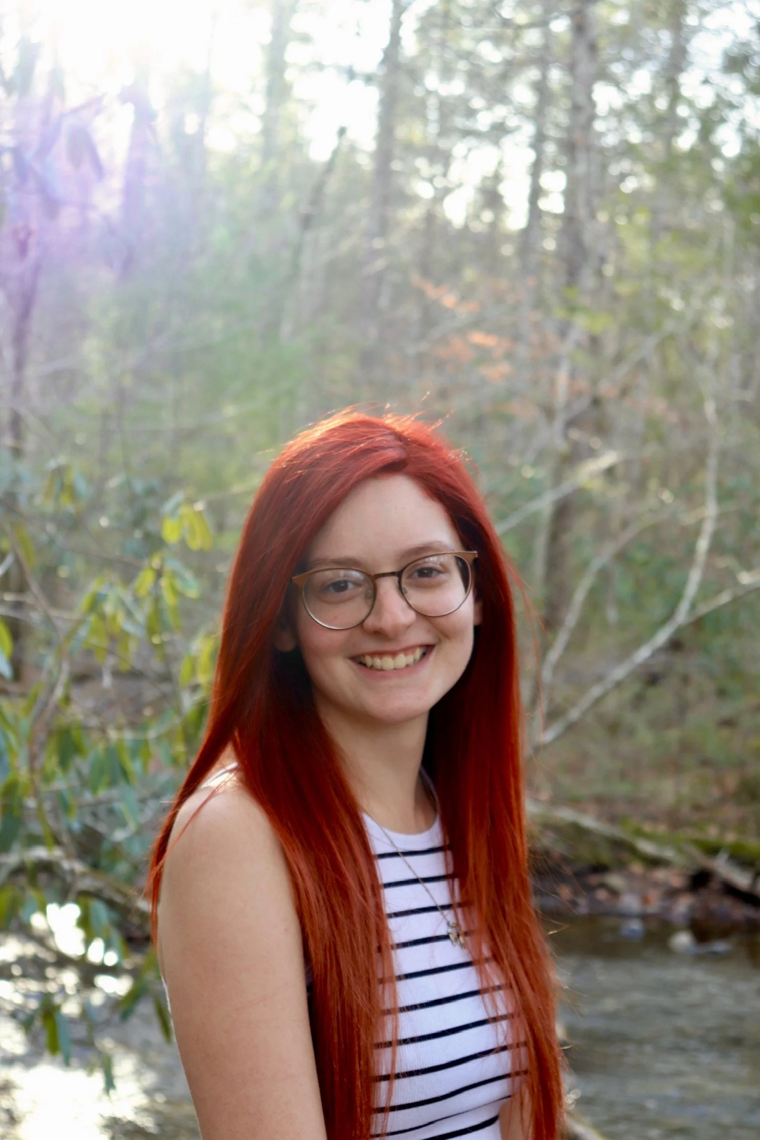 A smiling young woman with long red hair and glasses, wearing a striped top, standing outdoors near a stream in a forested area.