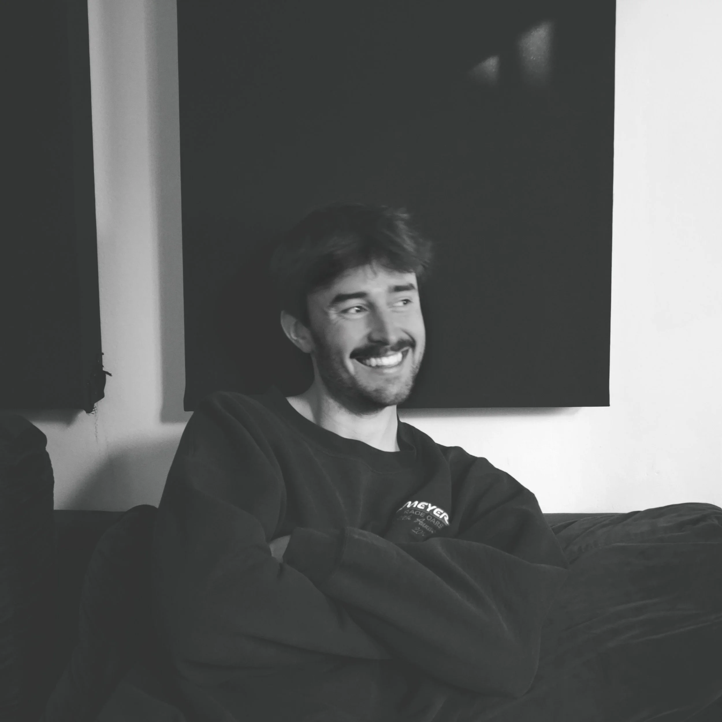 A black and white photo of a young man with short hair and a beard, smiling, wearing a dark sweatshirt with text on it. The background shows a large framed picture of himself smiling.