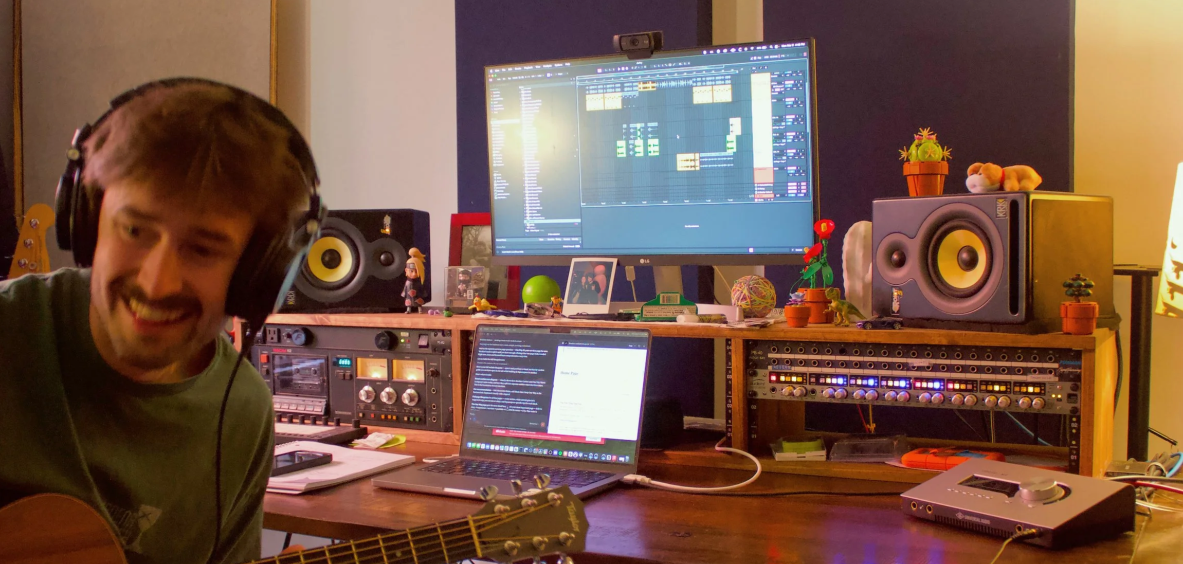 A person smiling and wearing headphones, sitting at a desk with a guitar, two computer monitors, and music equipment in a recording studio or home music workspace.