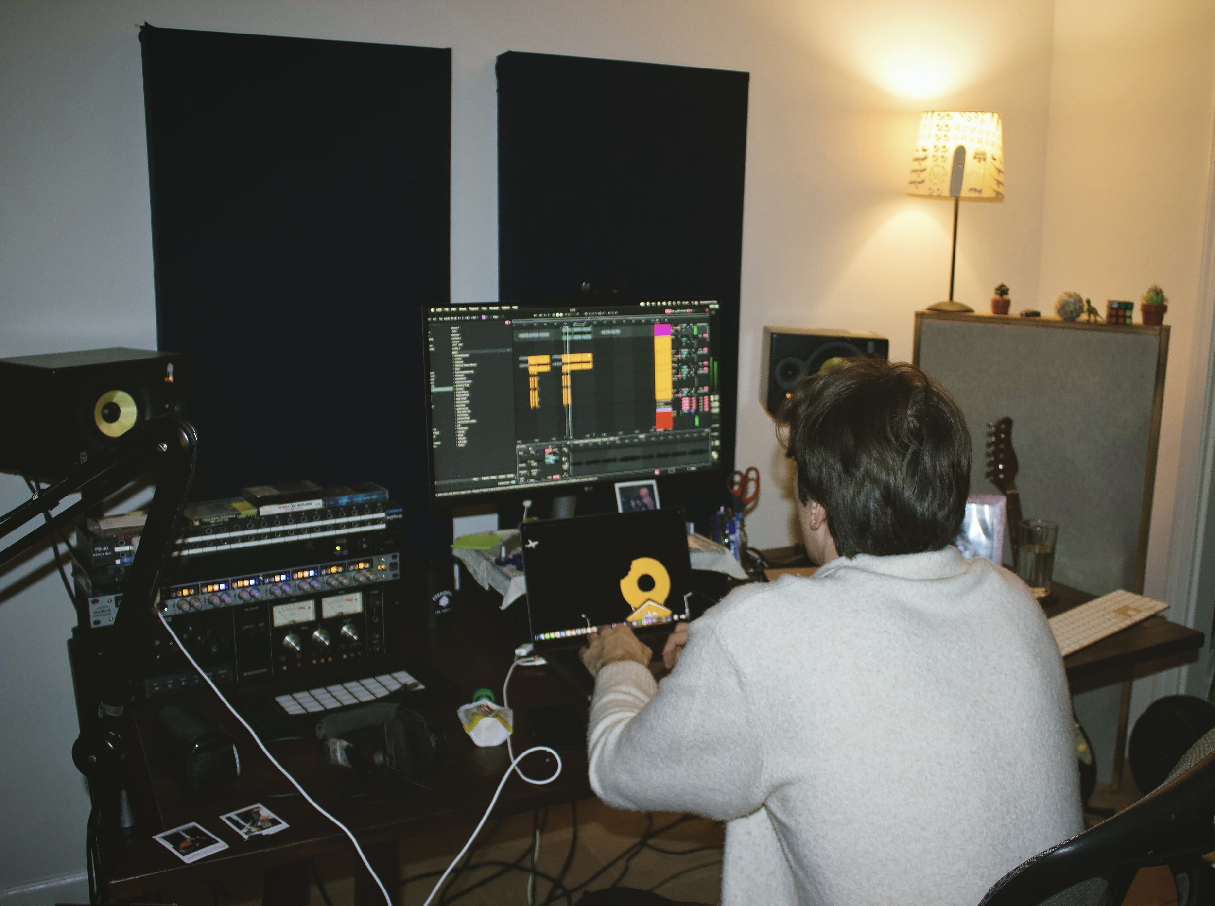 A person with short hair and glasses working on a computer with music production software in a home studio. The room has a lamp, potted plants, and a guitar in the background.