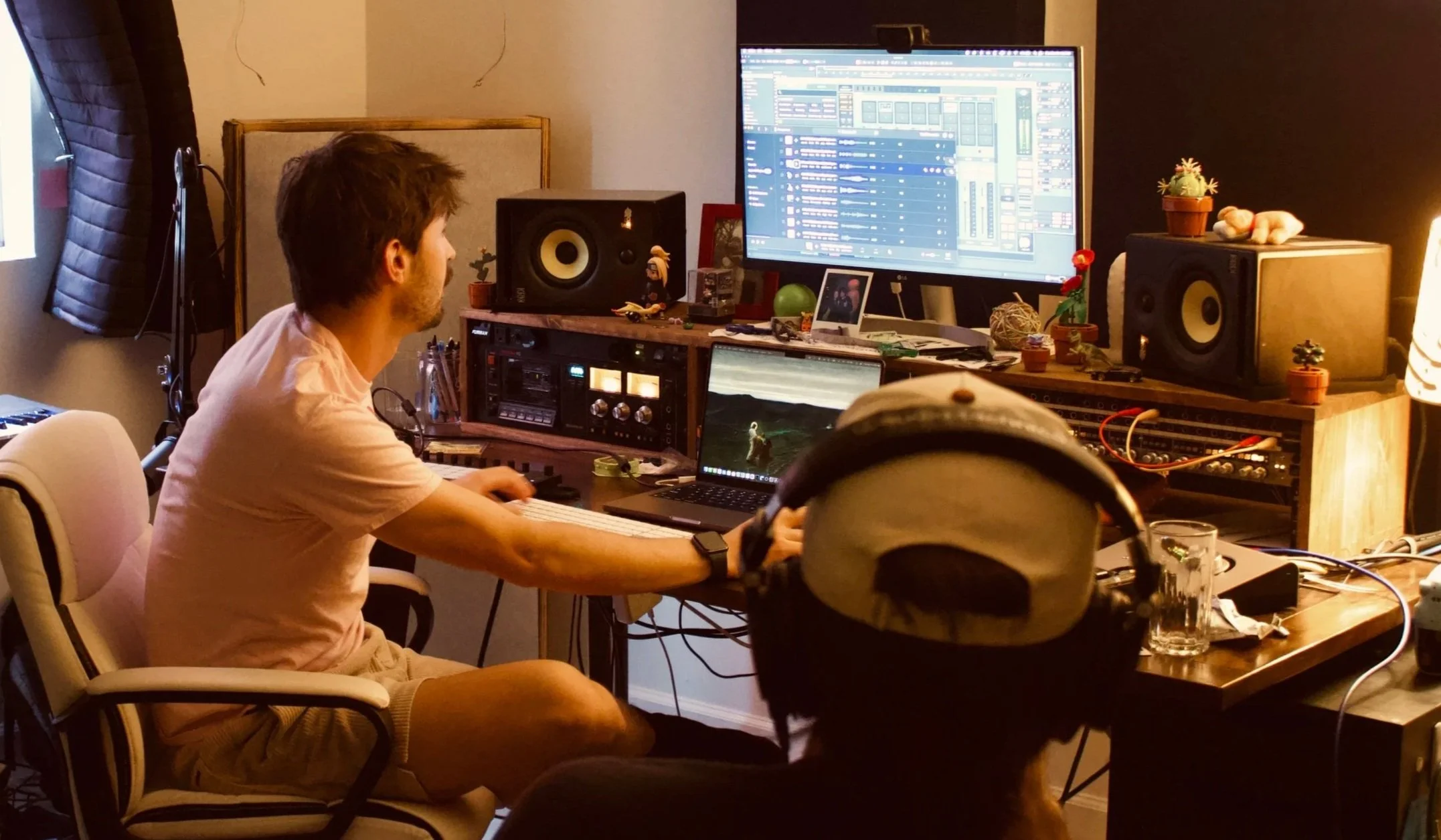 A man working on music production at a cluttered desk with multiple monitors, speakers, and audio equipment in a home studio, with small plants and decorative items around.