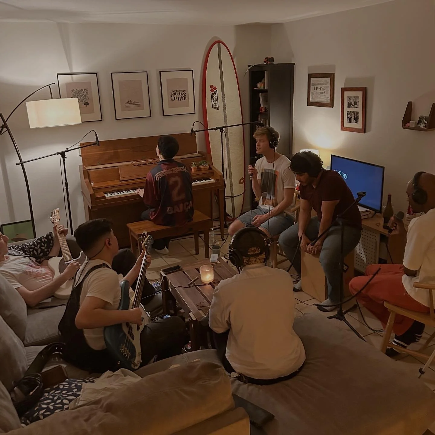 A group of people in a cozy living room recording a music session with instruments, microphones, and headphones, featuring a man playing piano, and others with guitars and singing.