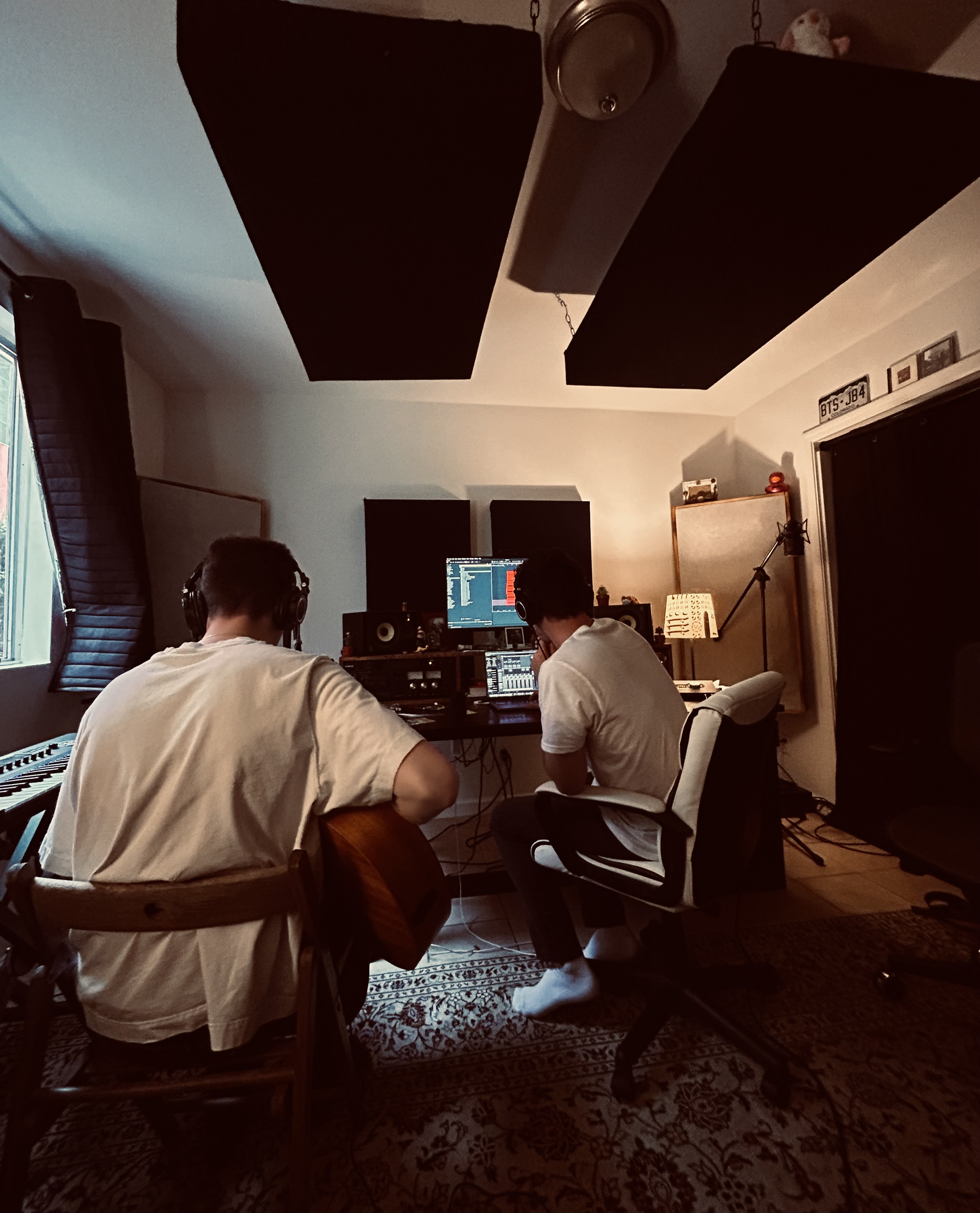 A recording studio with two men sitting at a desk with computers, wearing headphones. One man has white hair and is wearing a white shirt, while the other has dark hair and is also wearing a white shirt. There are soundproofing panels on the wall, a window with curtains, and a decorative area rug on the floor.