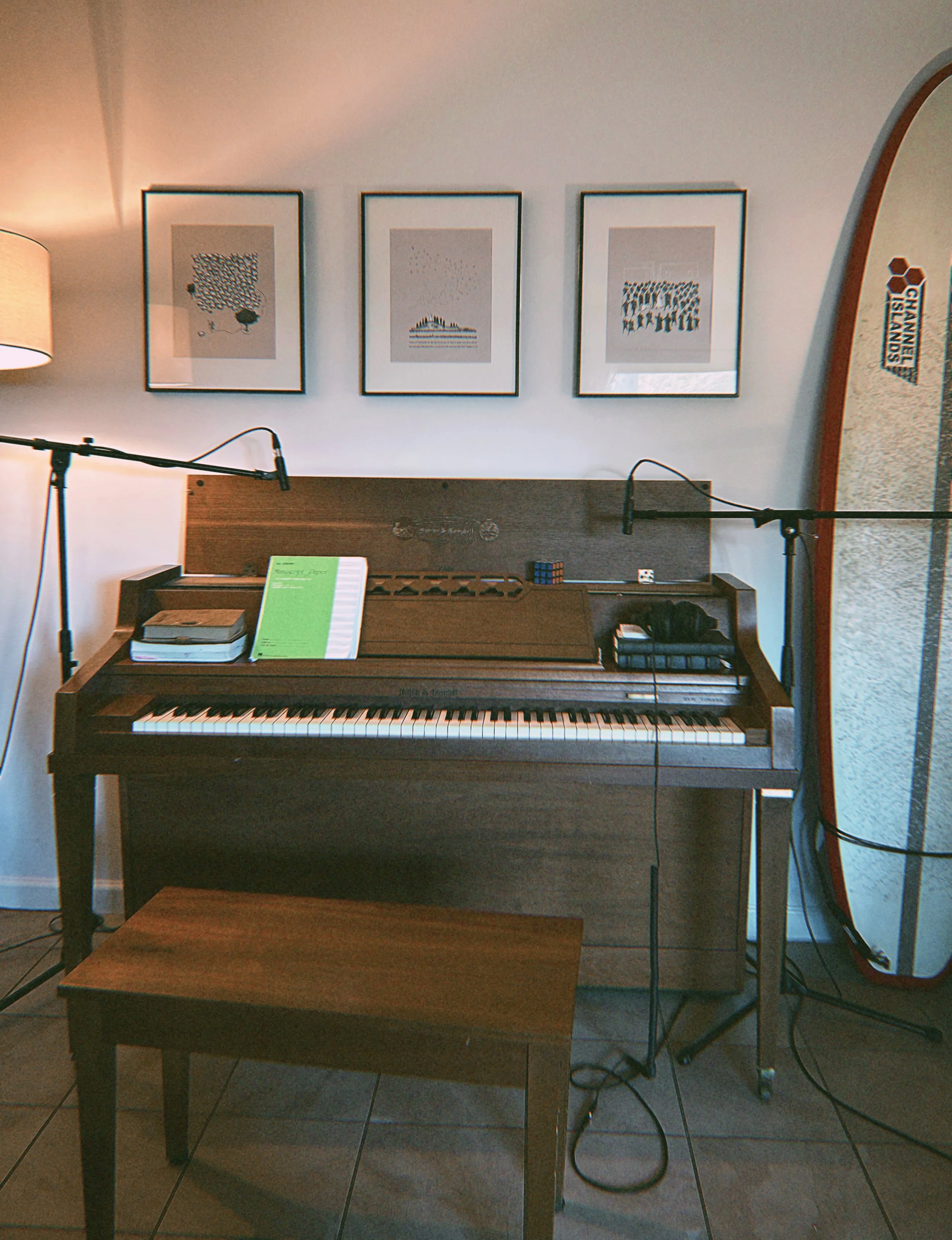 A piano with a book and sheet music on top, in a room with three framed abstract art pieces on the wall, two standing microphones, a traditional floor lamp, and a surfboard leaning against the wall.