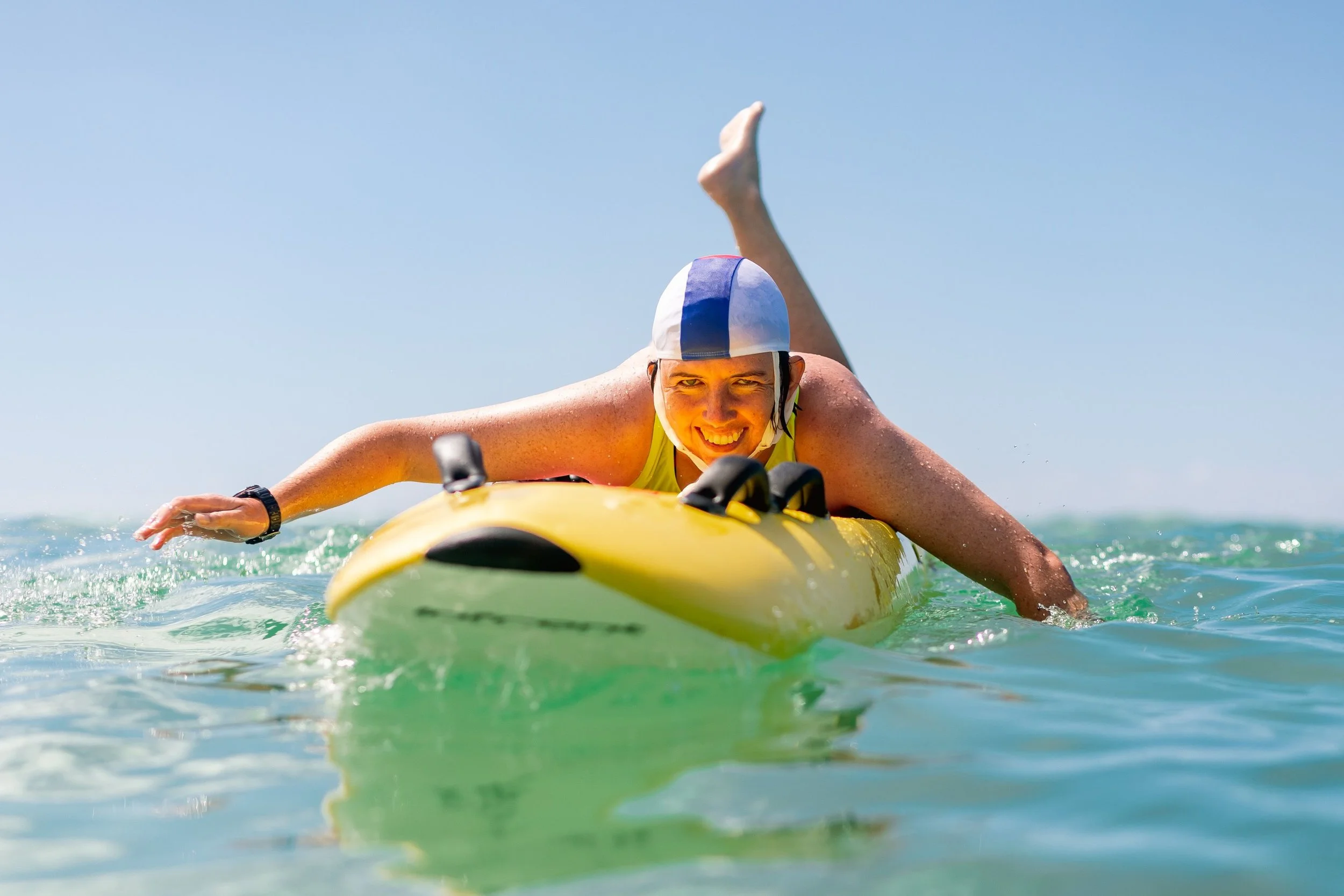 A person in a yellow tank top and white swim cap paddling on a yellow kayak in the ocean, smiling with a blue sky overhead.