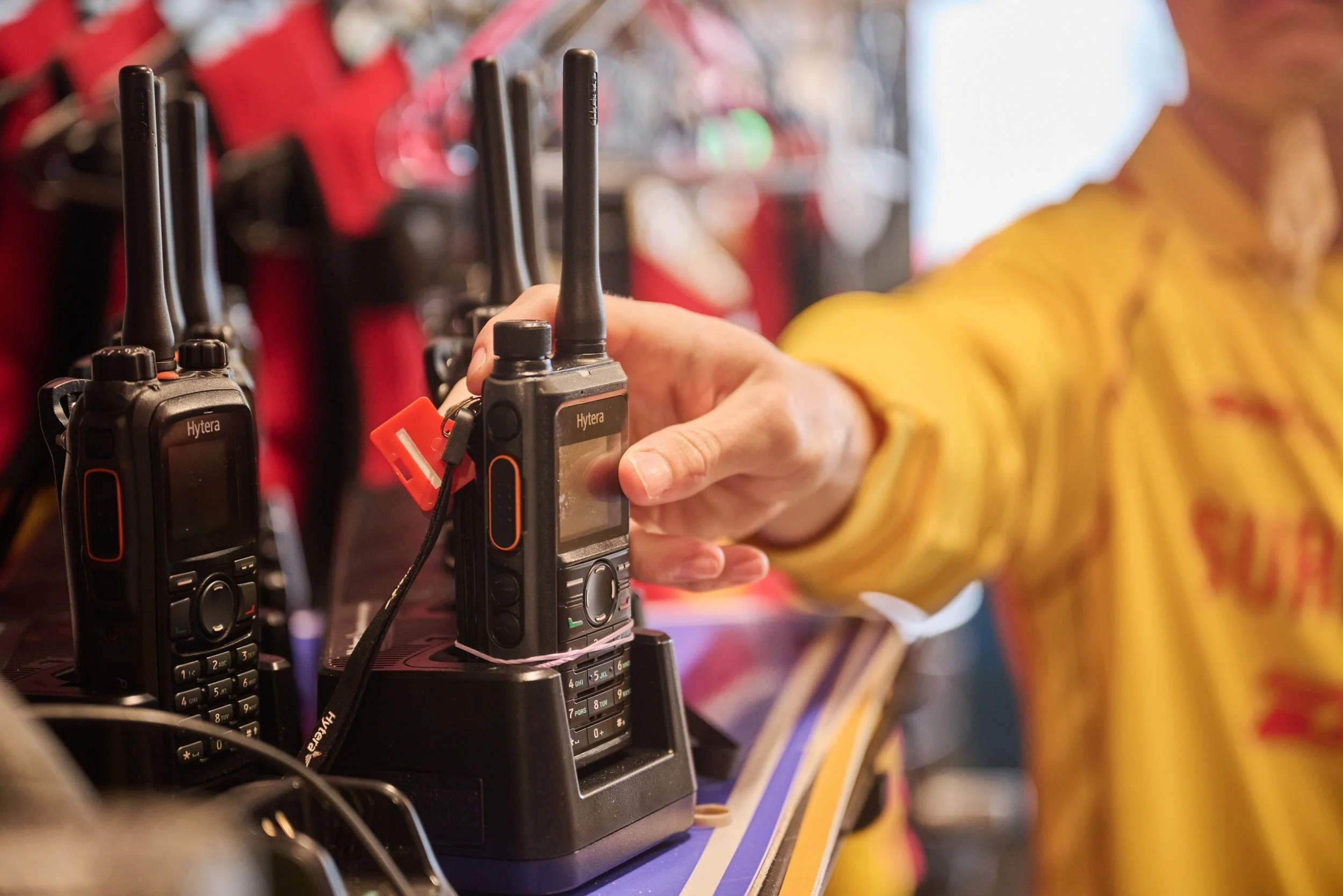 A person in a yellow top holding a walkie-talkie with rectangular screen, buttons, and antenna, surrounded by multiple similar walkie-talkies on a shelf.