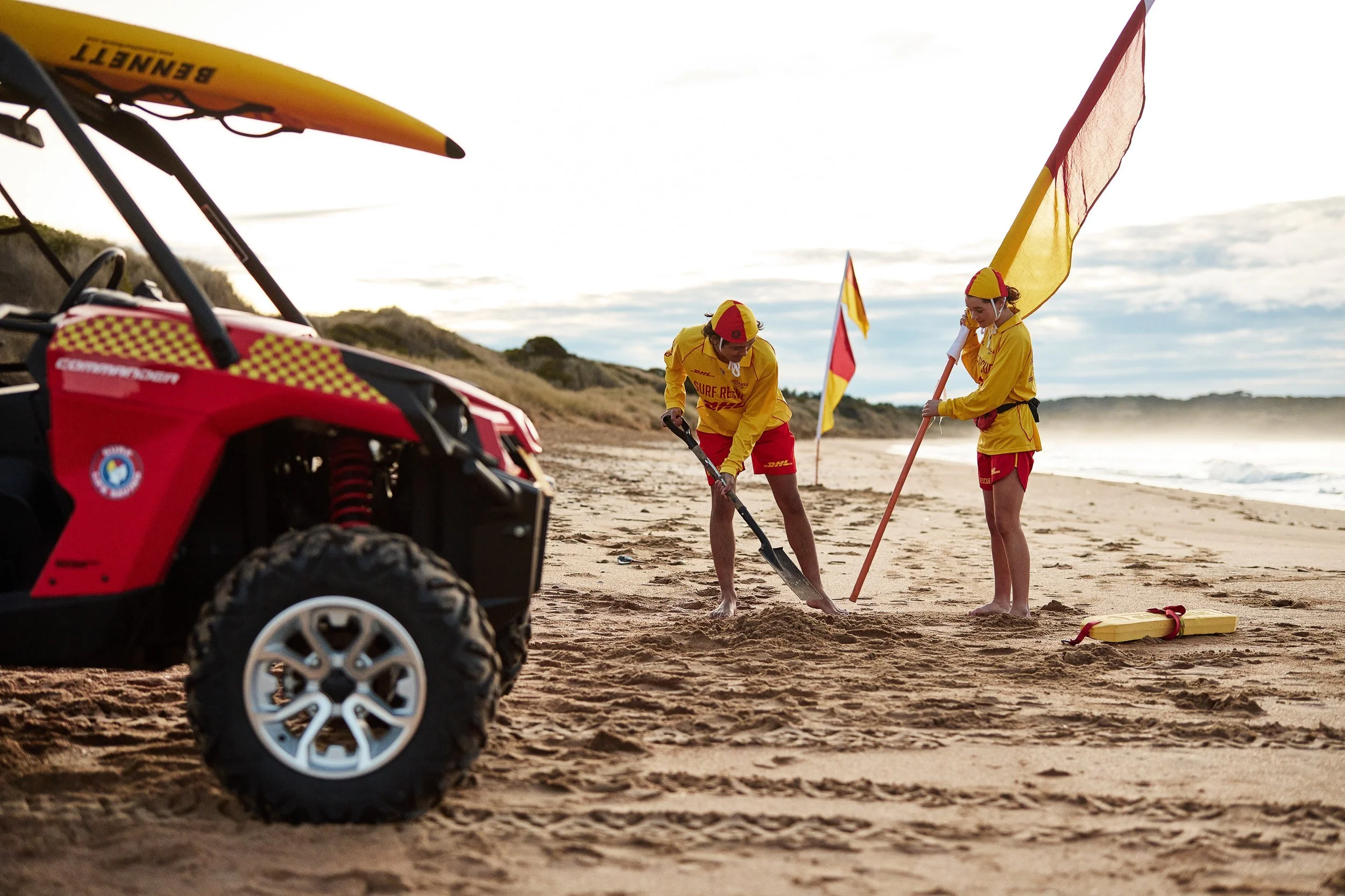 Two lifeguards on the beach, dressed in yellow and red uniforms, holding flags, with a rescue vehicle in the foreground, and the ocean and cloudy sky in the background.