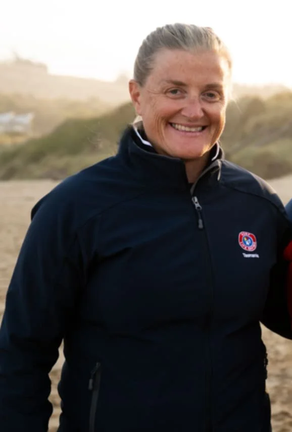 Smiling woman with light-colored hair tied back in a ponytail, wearing a dark jacket outdoors with a sandy or grassy landscape in the background.