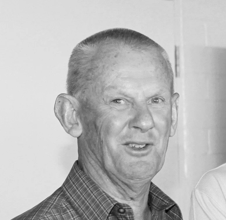 Black and white photograph of an elderly man with short, graying hair, wearing a checkered shirt, looking slightly to the side.